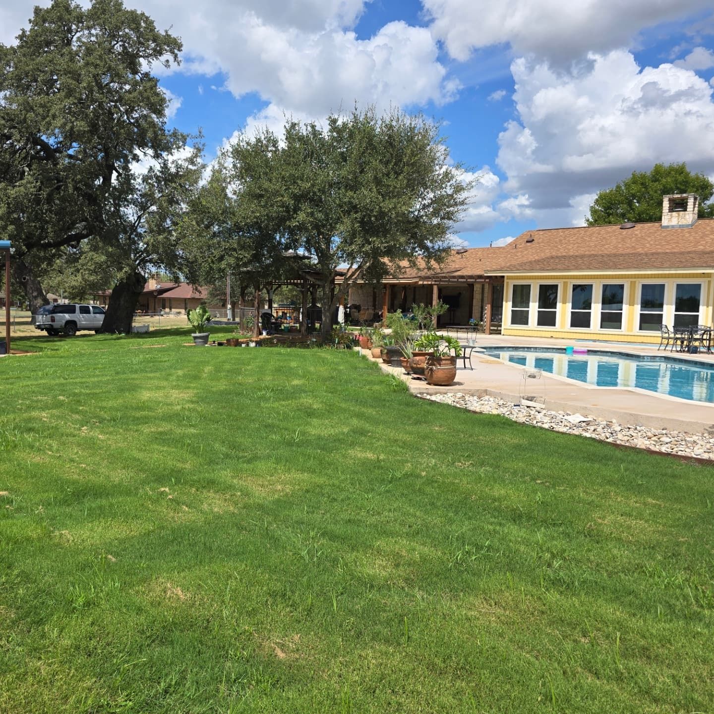 Green lawn, pool, and yellow house with blue sky and clouds.