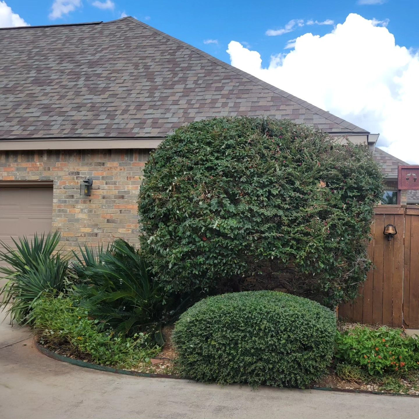 A house with a brown roof and a brick facade, green bushes, and a brown garage door.