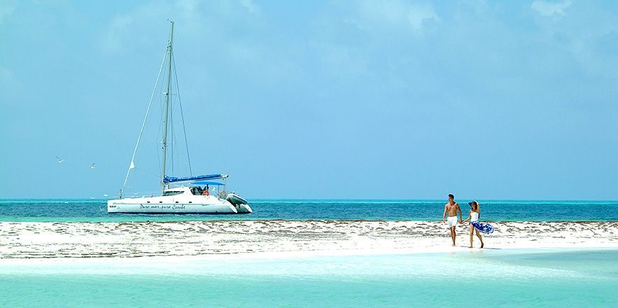 Una pareja parada en una playa con un velero al fondo.