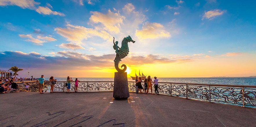 Un grupo de personas están de pie alrededor de una estatua de un caballito de mar al atardecer.