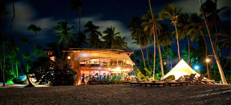 Una casa grande se ilumina por la noche en una playa rodeada de palmeras.