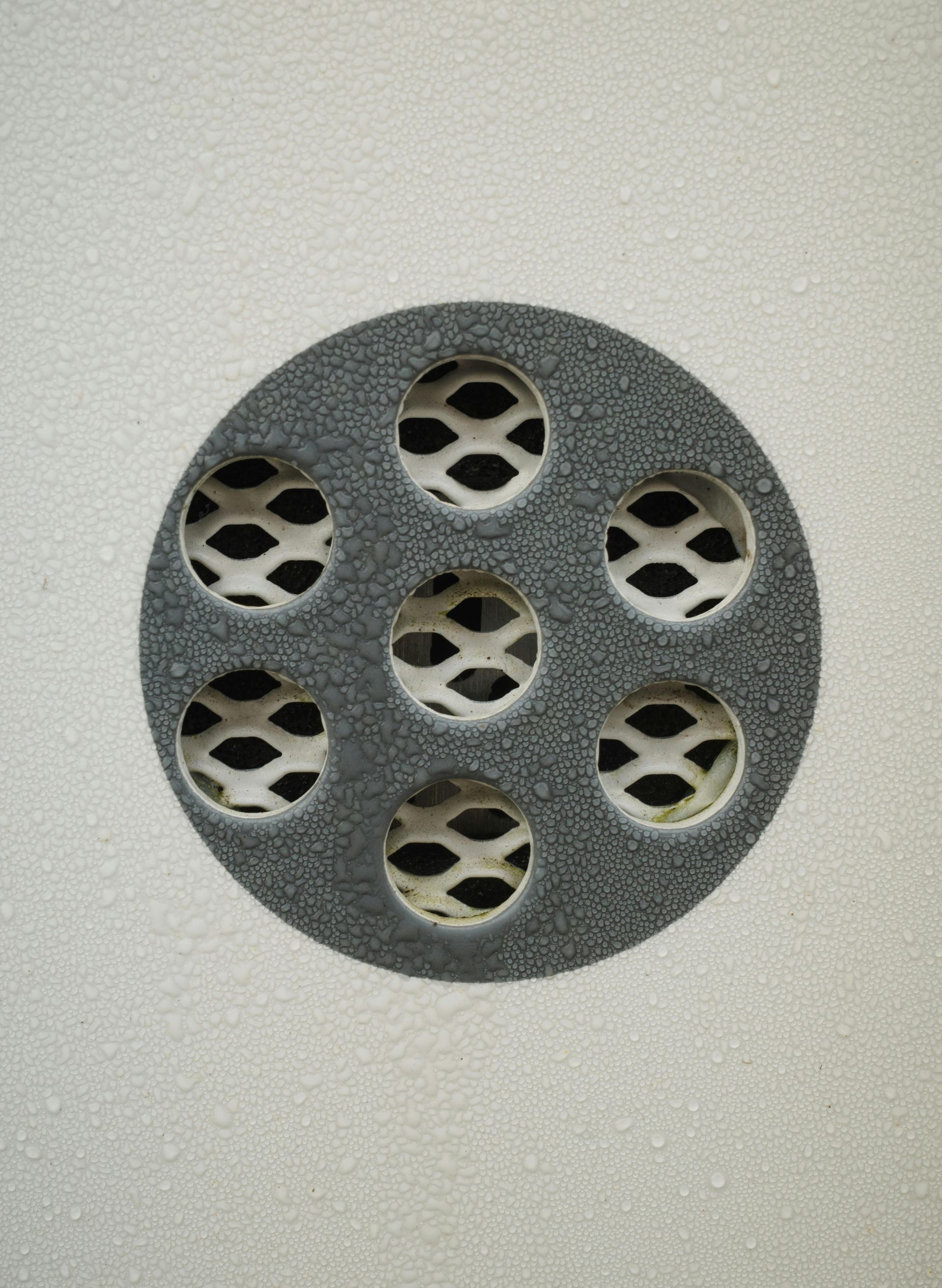 Circular vent with seven holes on a white foam surface. The vent is dark grey with a white, honeycomb-like pattern visible inside.