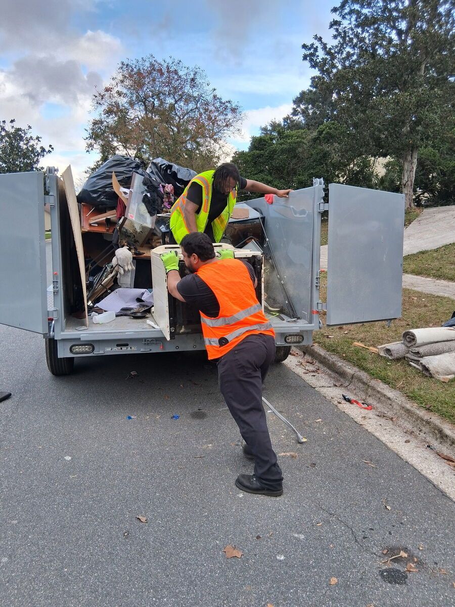 Two workers loading trash into a truck on a residential street; one in an orange vest, one in yellow.
