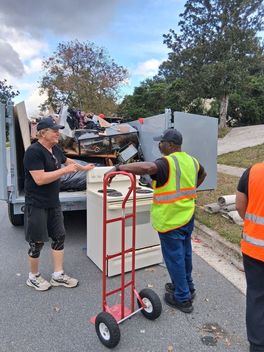 Two men beside a trash truck, discussing. One wearing a neon vest, the other a cap and knee brace. A dolly and discarded items are present.