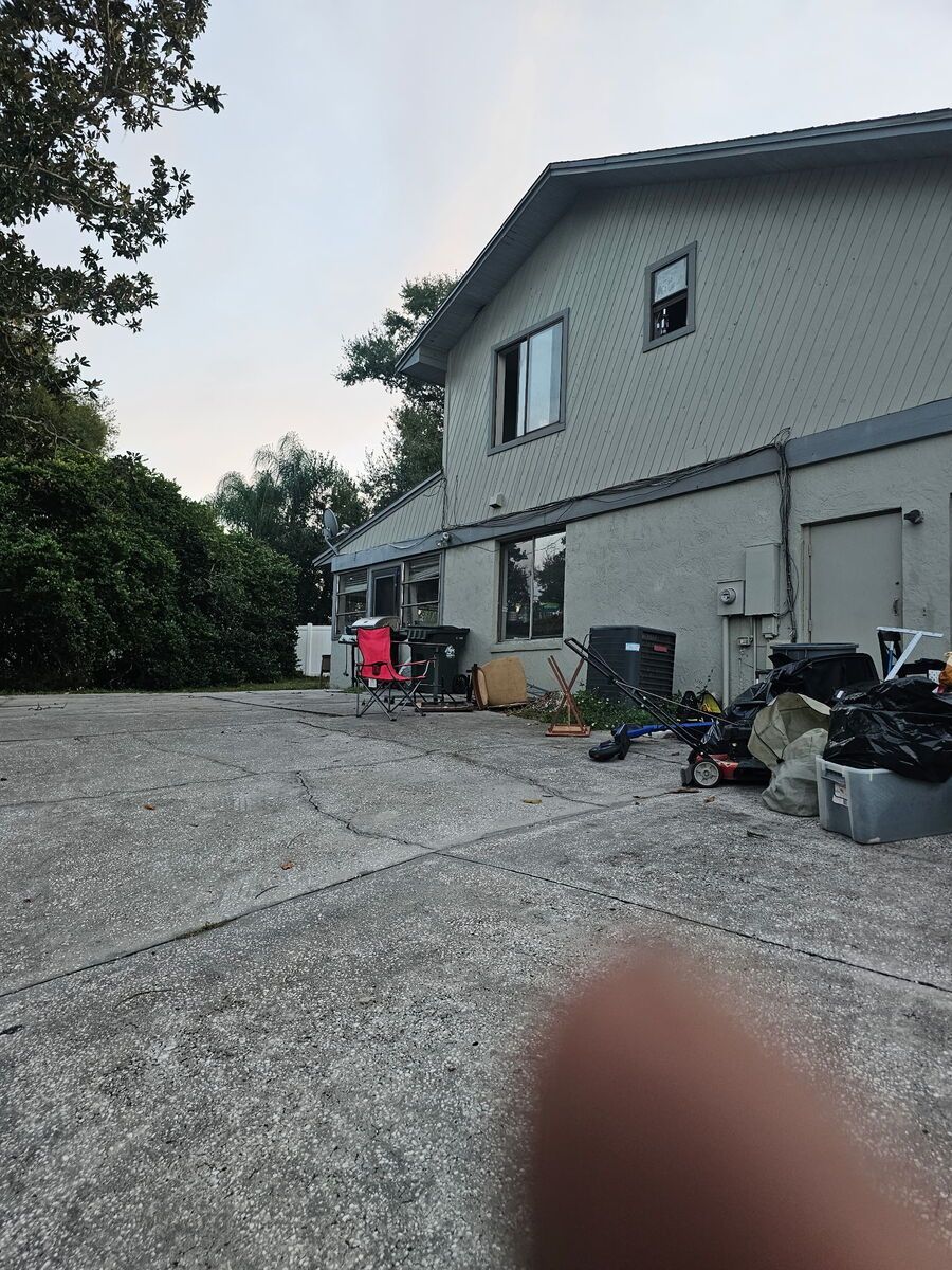 Back of a two-story gray house with a cracked concrete yard. Trash cans and debris are visible.
