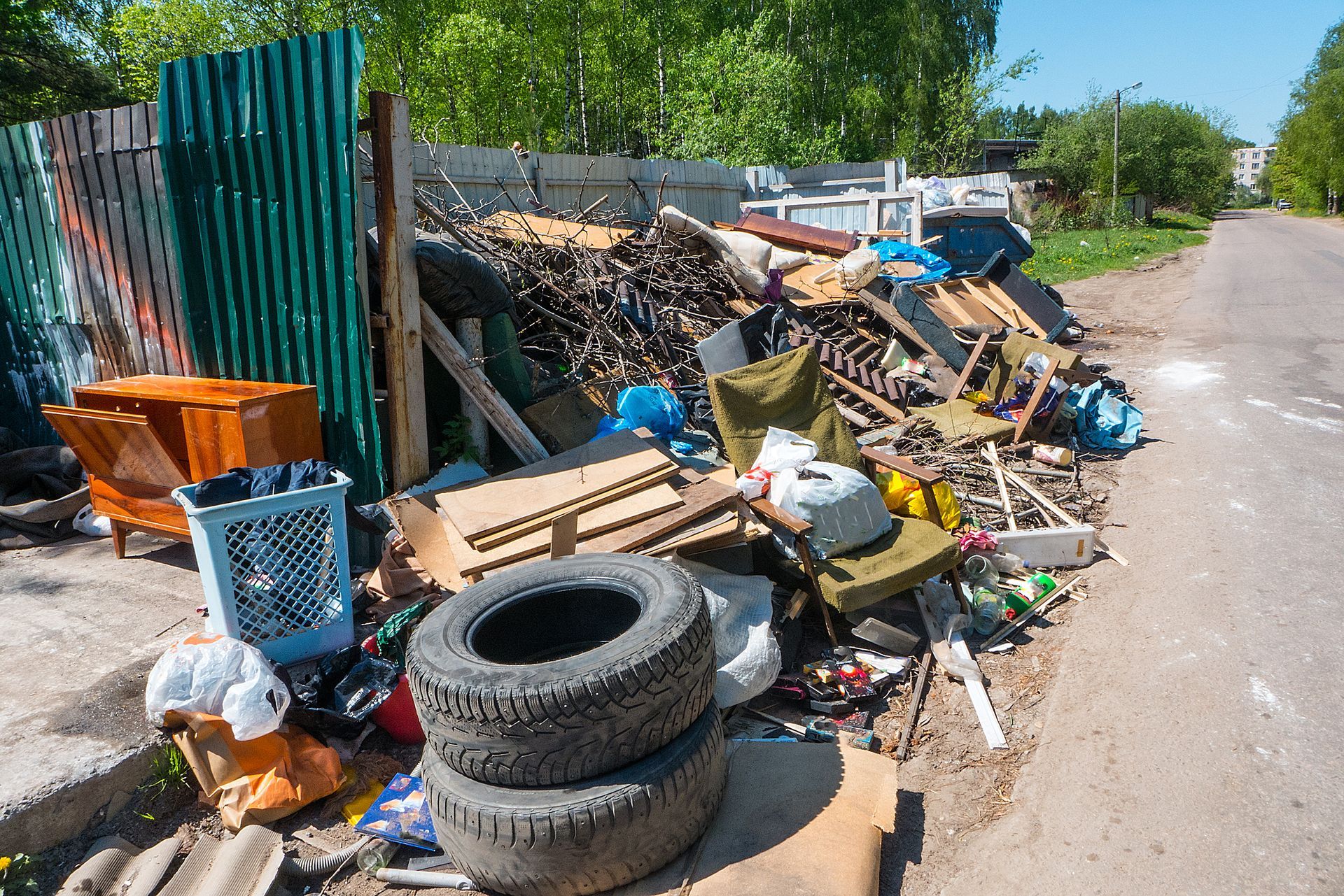 Pile of trash and debris on the side of a road, including tires, furniture, and wood, next to a metal fence.