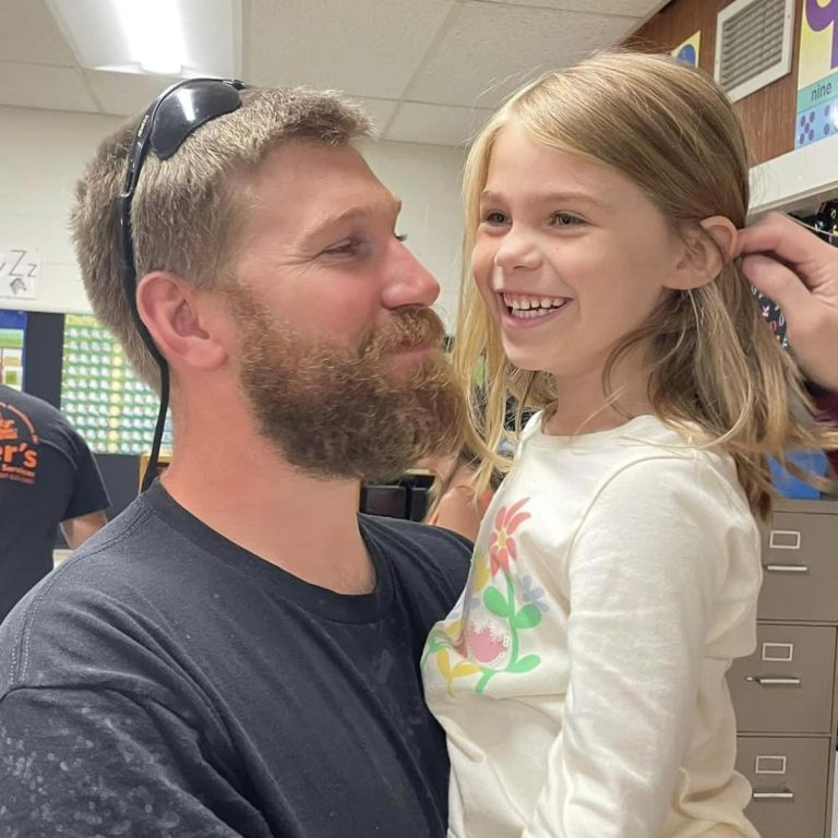 A bearded person holds a smiling child whose hair is being gently tucked behind their ear in a classroom setting.