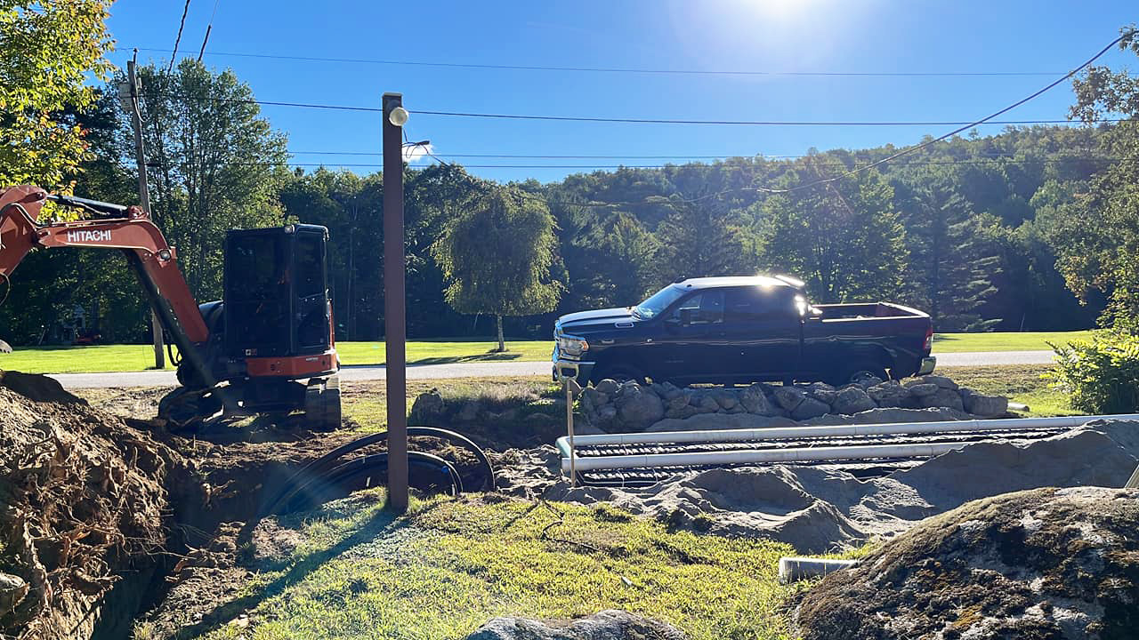 An excavator works on a construction site next to a dark pickup truck with a line of pipes laid on the ground.