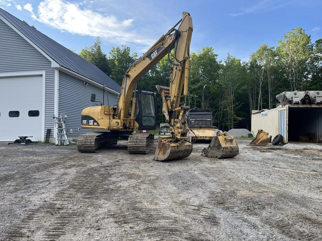 A large yellow Caterpillar excavator parked on a gravel lot next to a grey building and a shipping container.