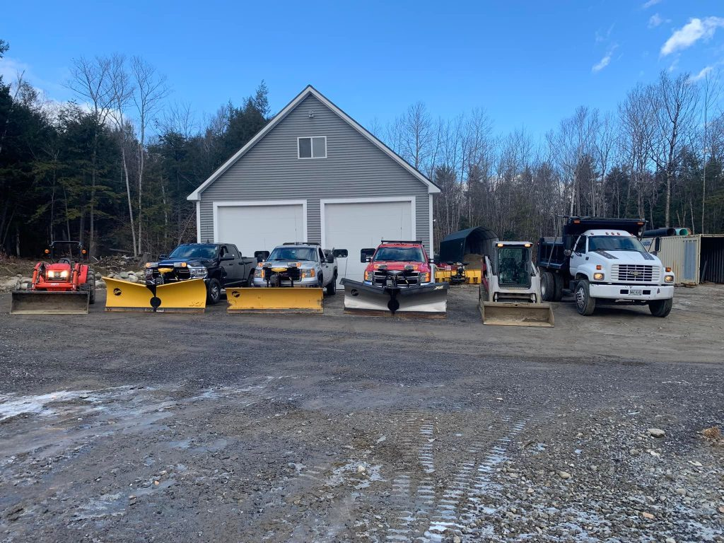 A line of trucks and equipment with snowplows parked in front of a gray garage on a gravel lot under a clear blue sky.