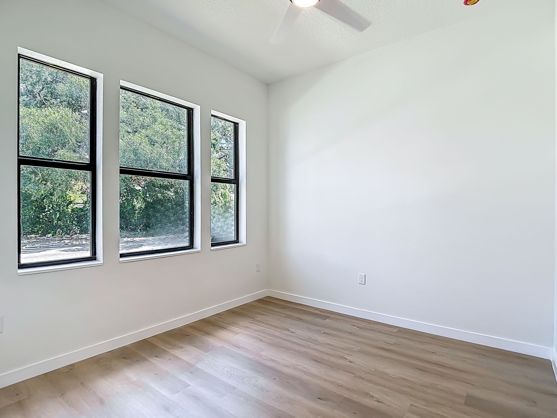 Empty room with three windows and wooden floors; white walls and a ceiling fan.