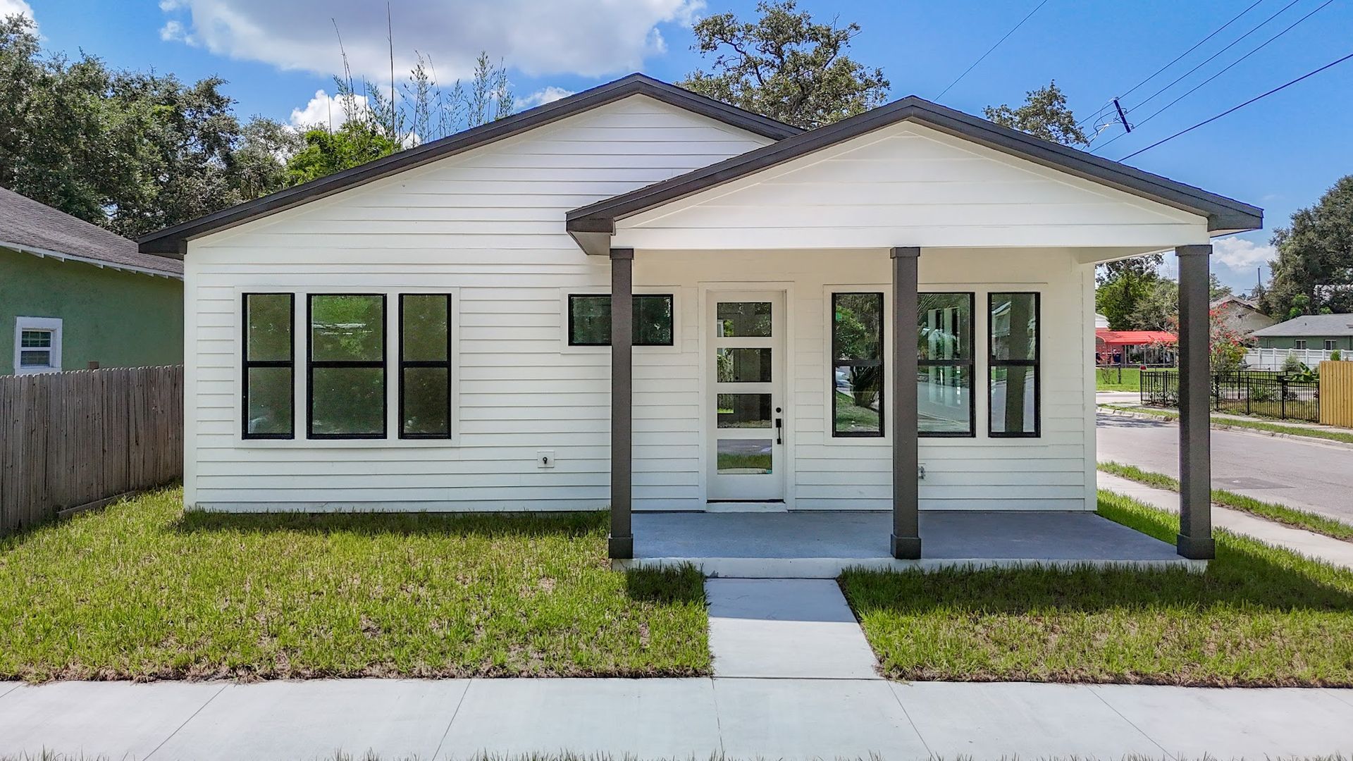 White house with dark trim and porch, green lawn, sidewalk in front.