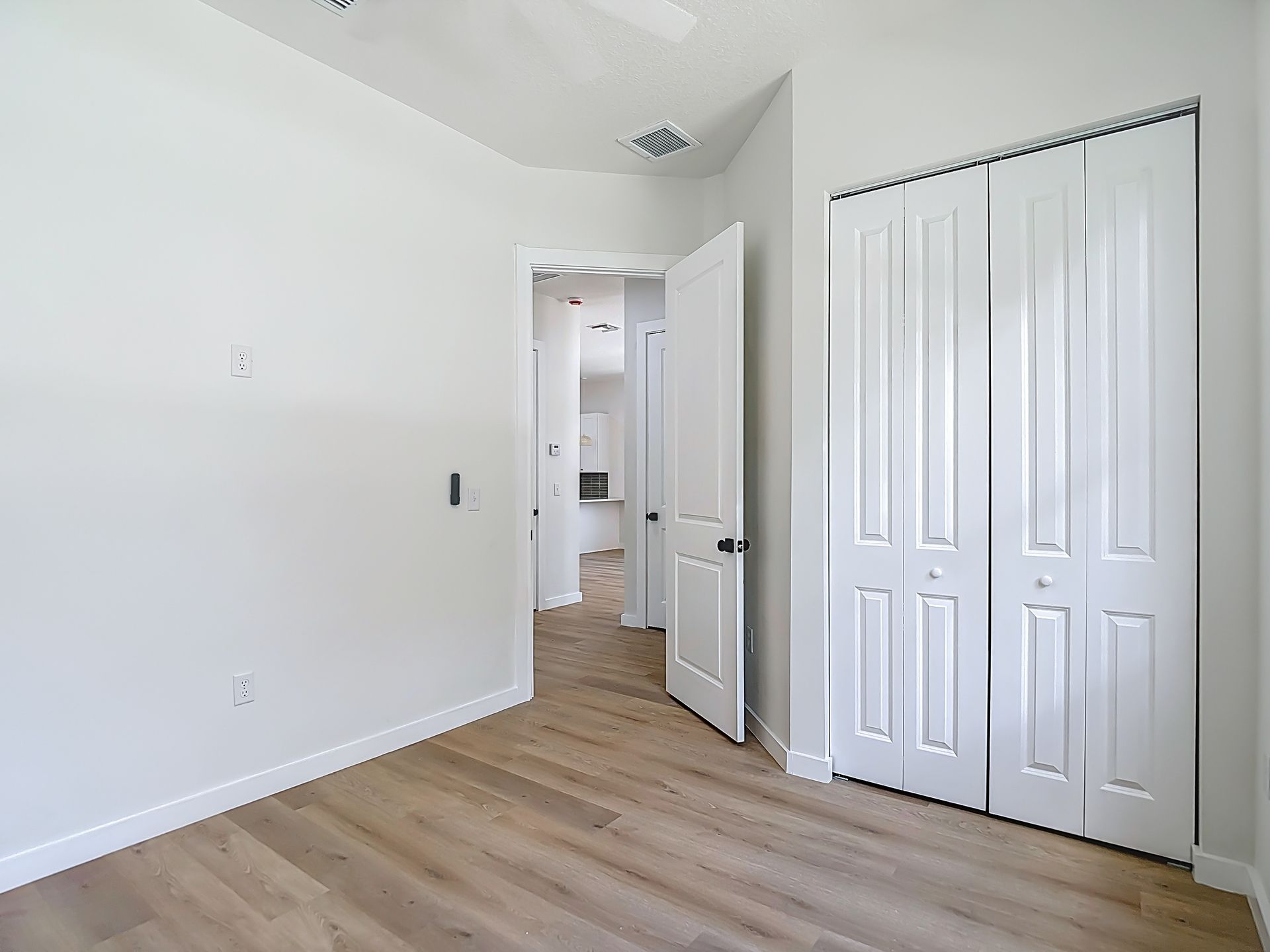 Empty bedroom with a white closet door, wooden floor, and an open door to a hallway.