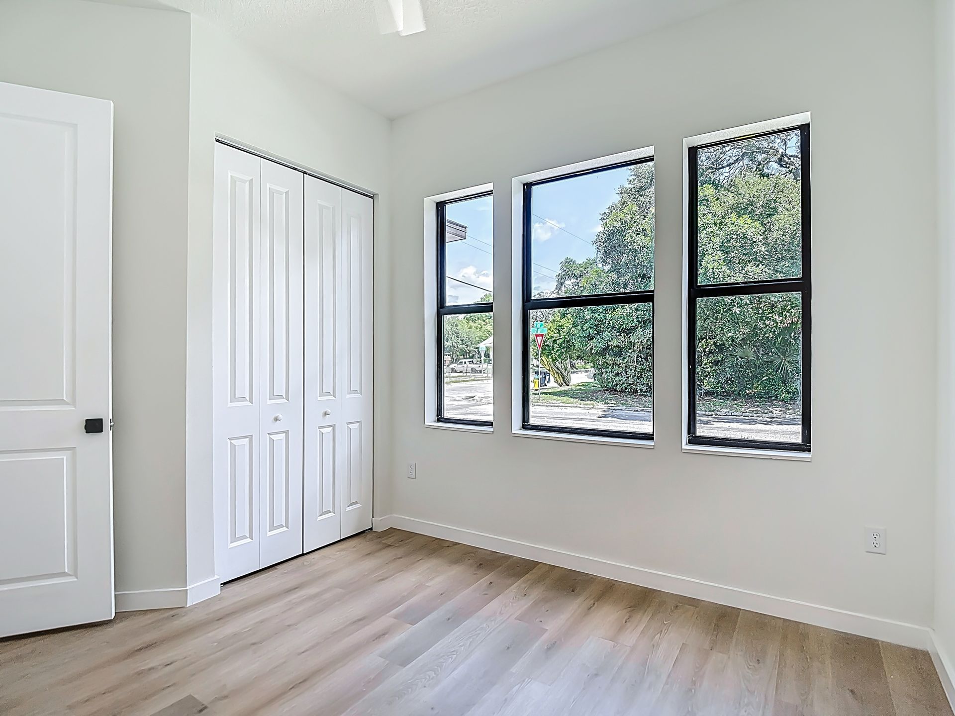 Empty bedroom with wood floors, closet, door, and three windows overlooking trees.