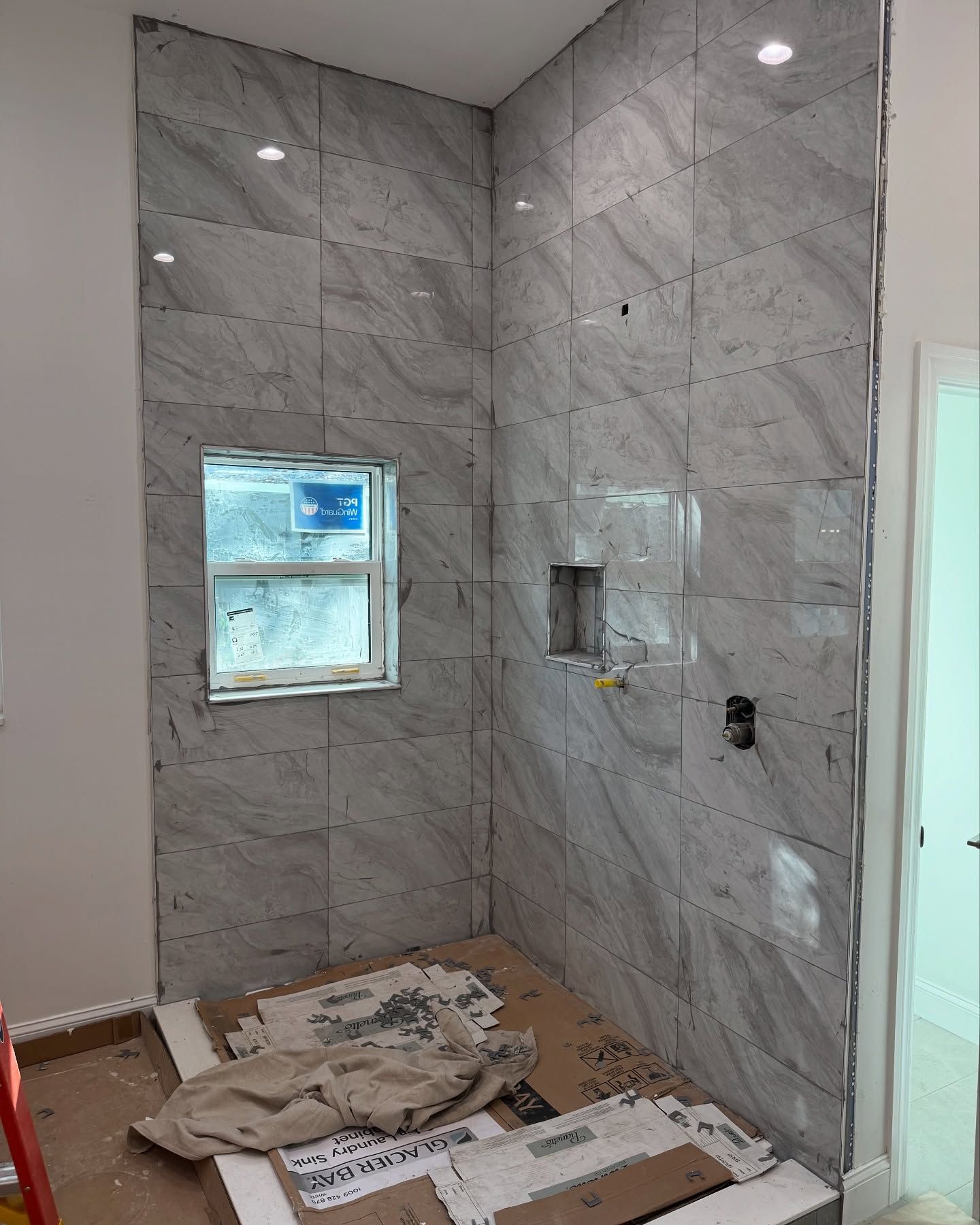 Shower stall under construction, tiled walls in a light gray marble pattern. Window and recessed niche visible.