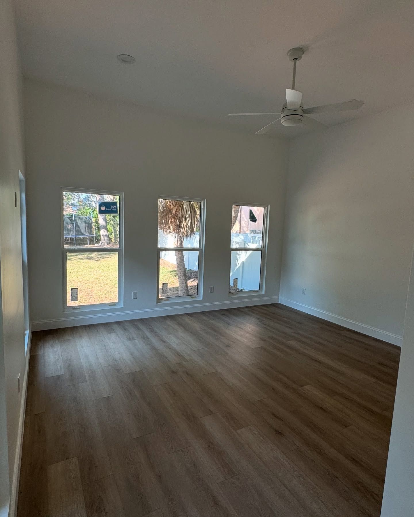 Empty room with wooden floor, three windows, white walls, and a ceiling fan.