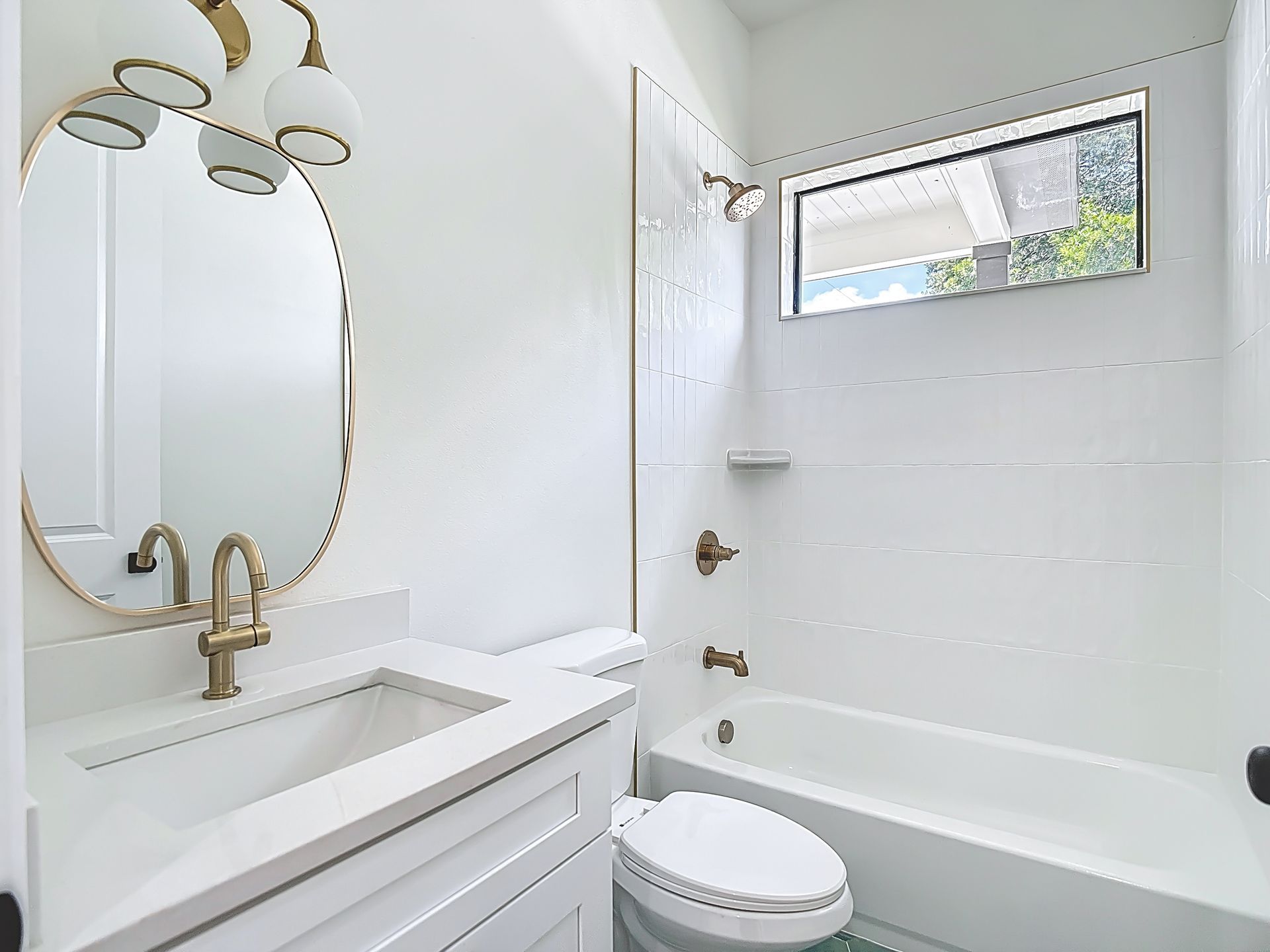 White bathroom with gold fixtures, oval mirror, and bathtub.