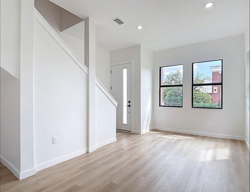 Empty, light-filled room with hardwood floors, a staircase, and windows. White walls and door.