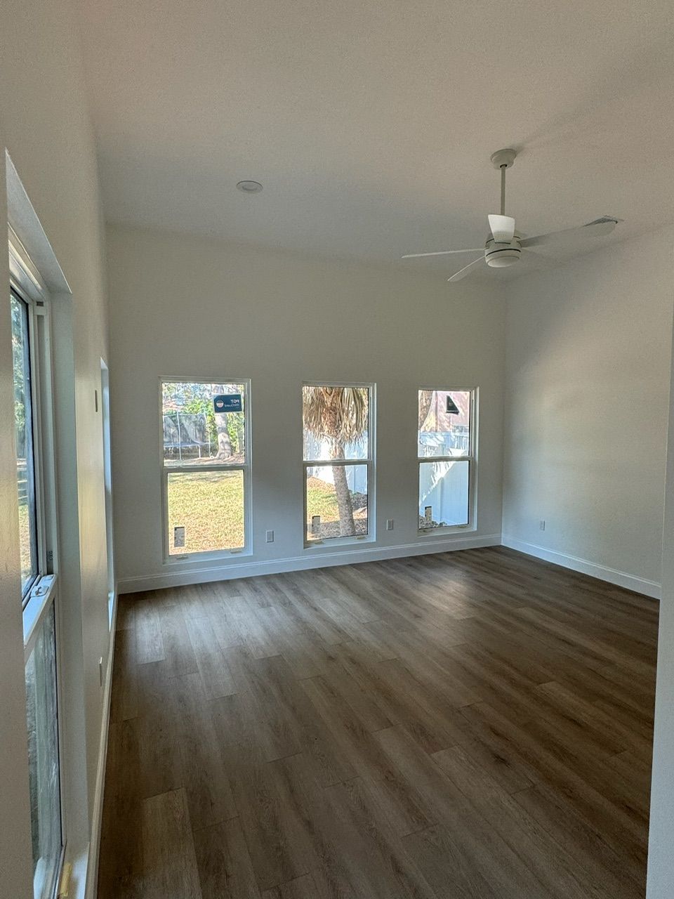 Empty living room with wood-look flooring, multiple windows, high ceiling, and ceiling fan.