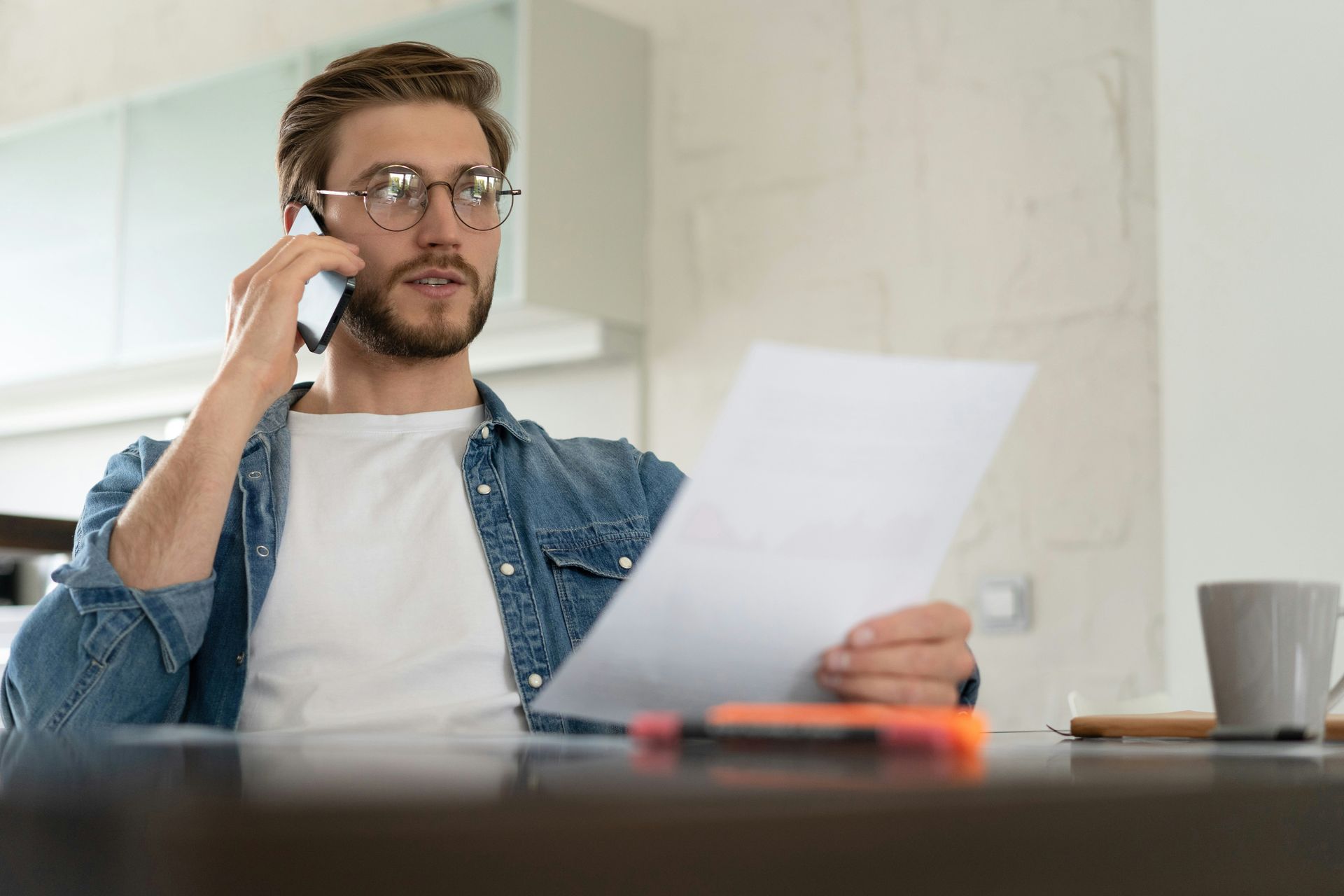 A man is sitting at a table talking on a cell phone while holding a piece of paper.