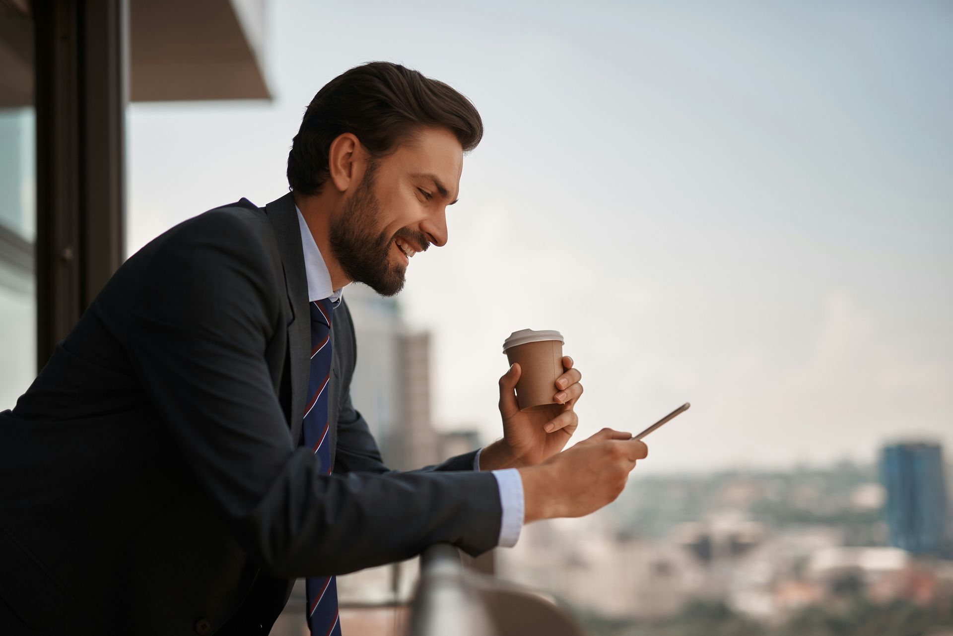 A man in a suit and tie is sitting on a balcony holding a cup of coffee and looking at his phone.