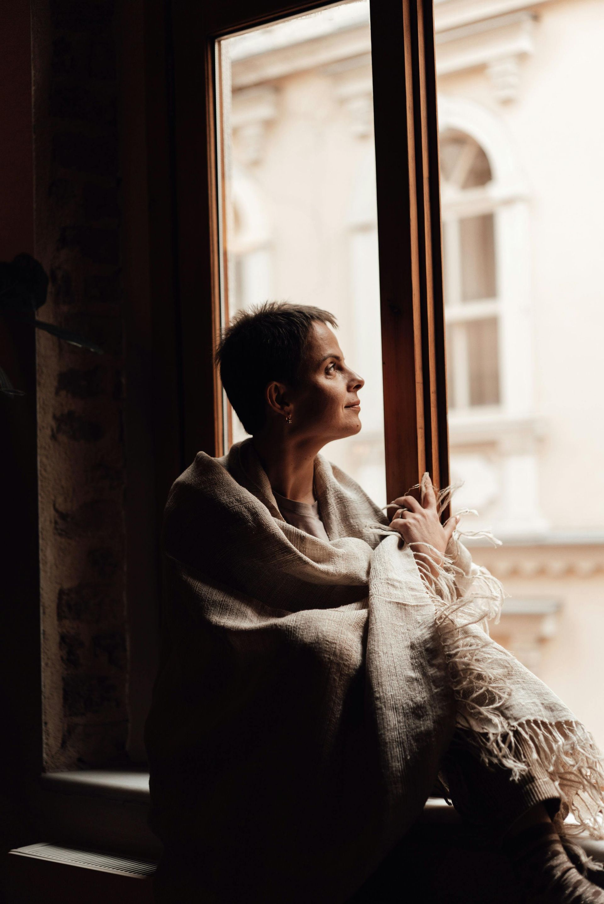 A woman wrapped in a blanket is sitting on a window sill looking out the window.