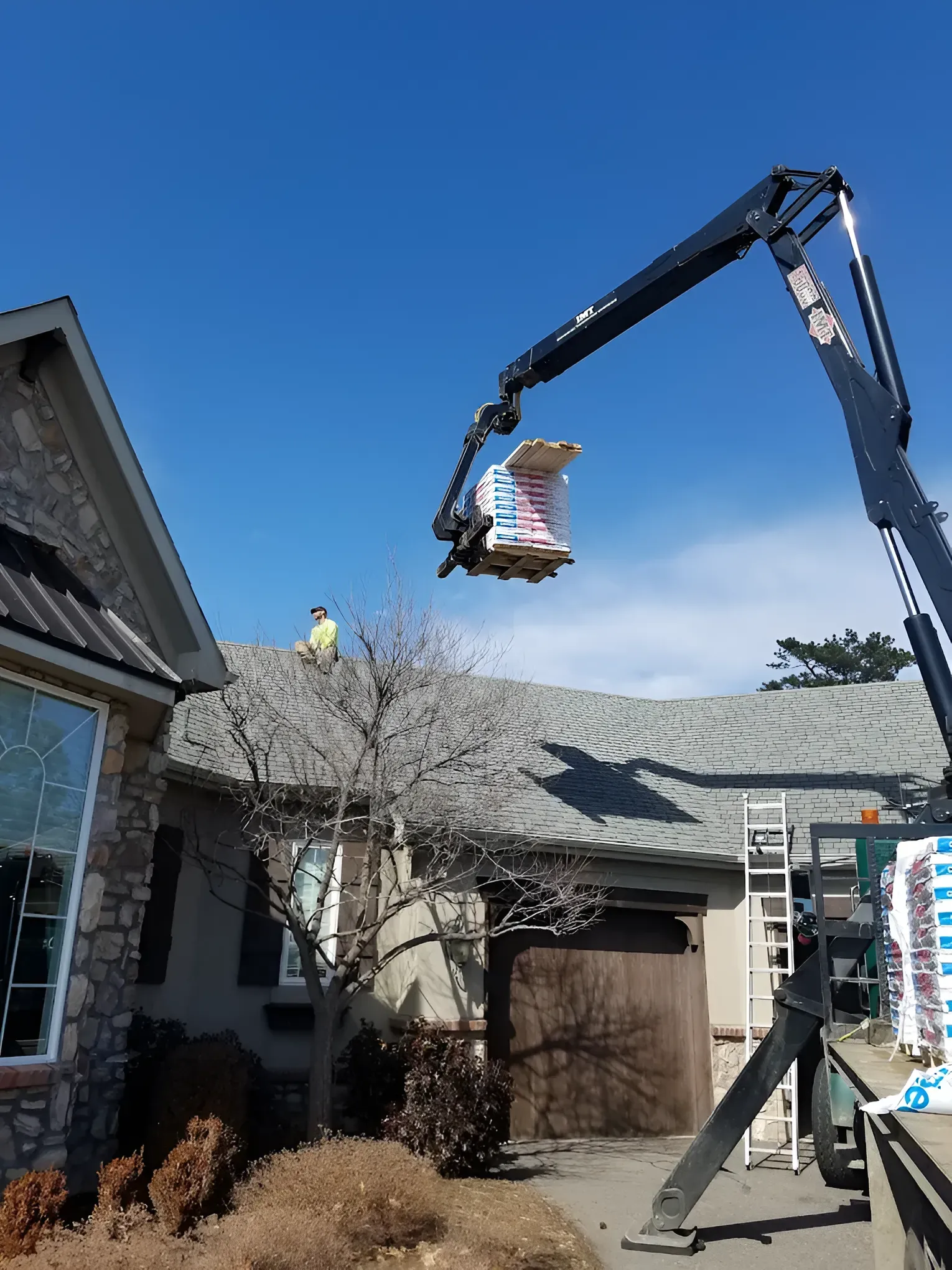 Crane lifting shingles onto a house roof; worker on roof; clear, sunny sky.