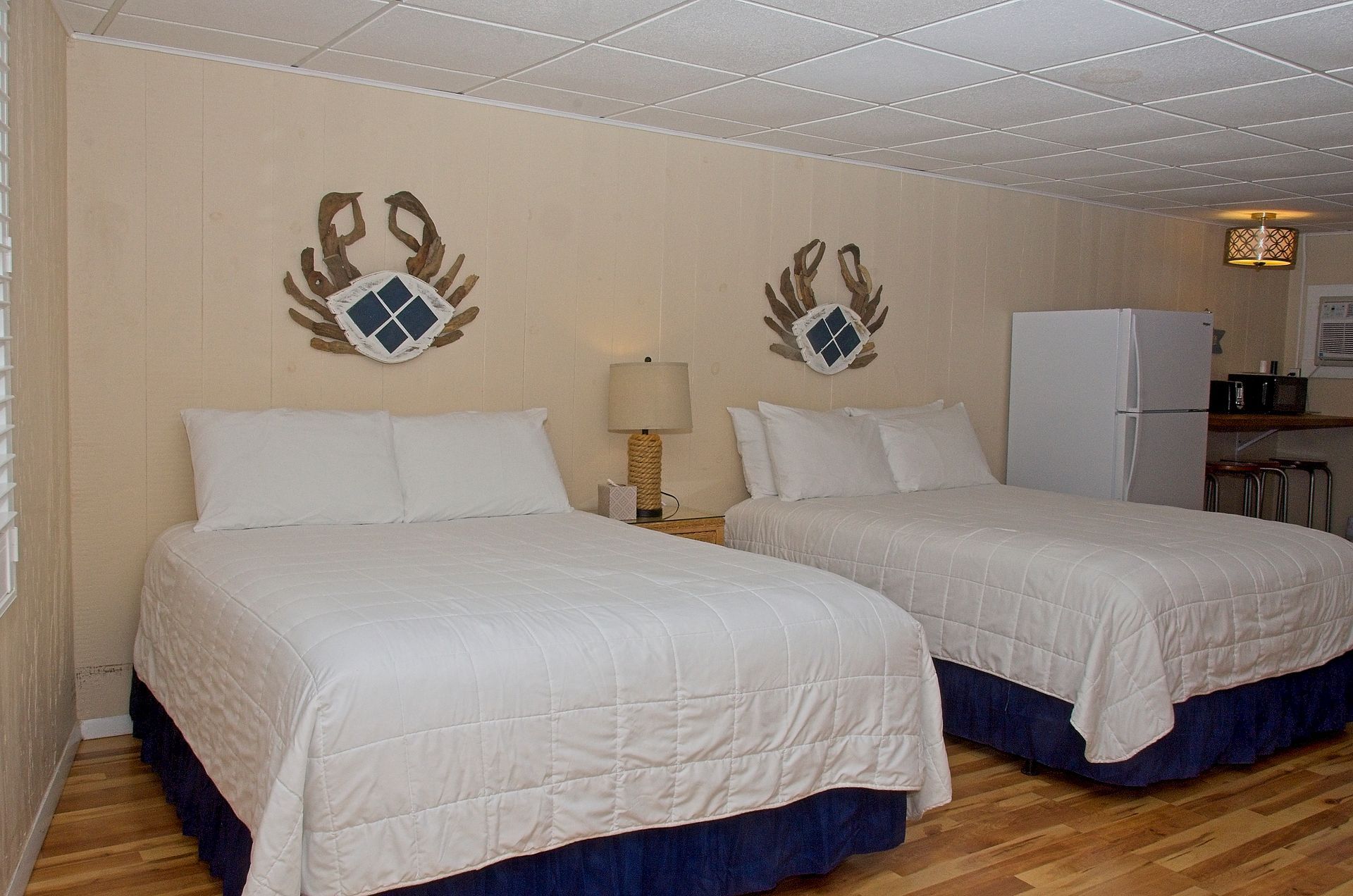 A motel room with two queen beds, crab art, and a small refrigerator. The room has beige walls and a light wood floor.