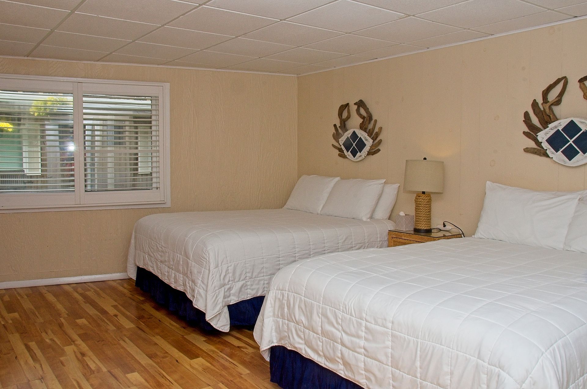 A hotel room with two queen beds, a window with blinds, and crab art on the walls. The room is beige with light wood floors.