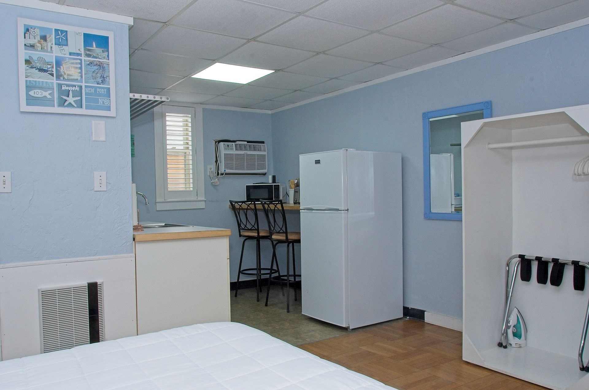Small, light blue studio apartment with a bed, refrigerator, and small dining area. Artwork with beach themes hangs on a wall.