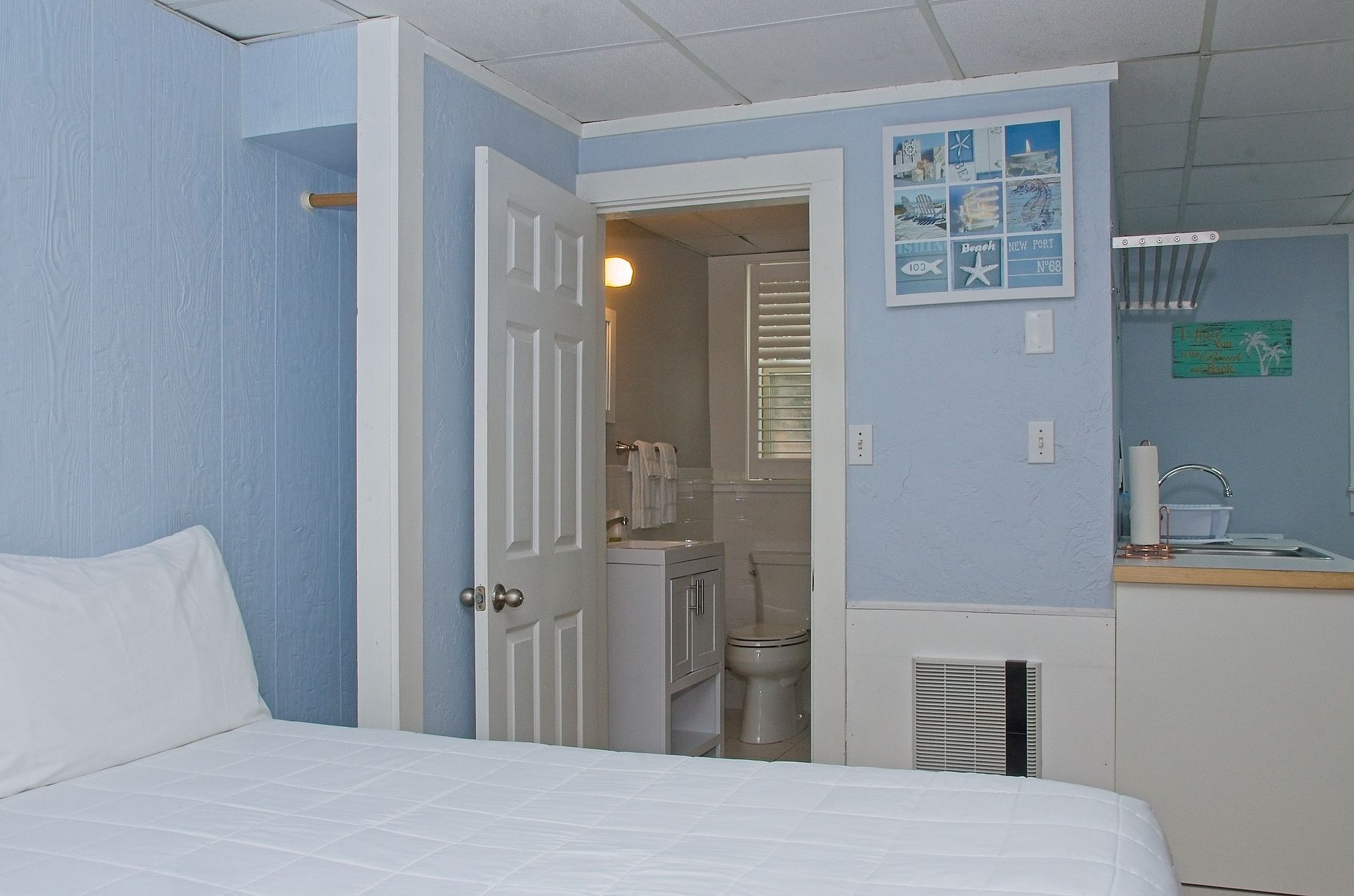 A small, light blue room with a bed, an open doorway to a bathroom, and decorative art.