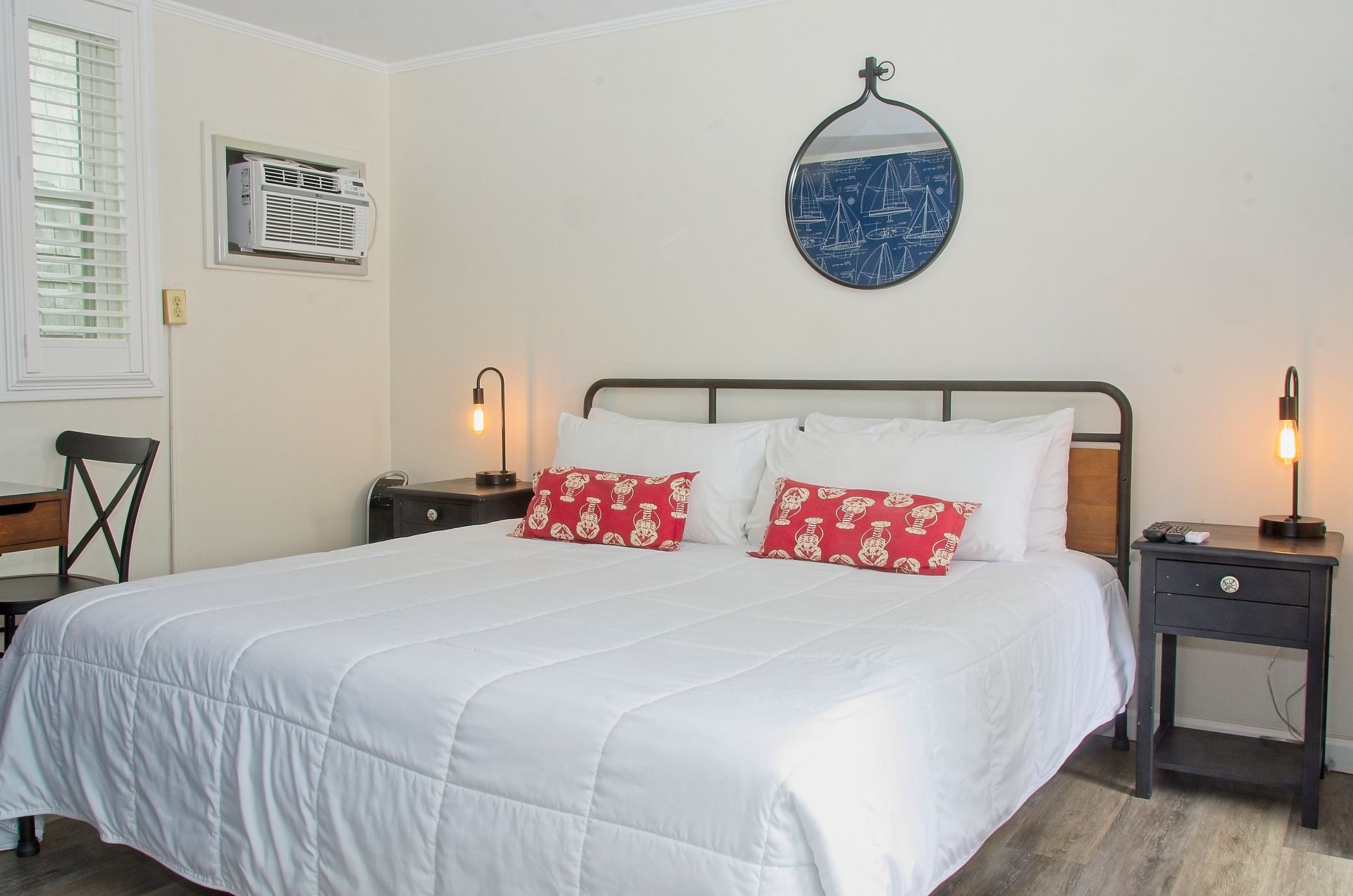 A hotel bedroom with a queen bed, nightstands, and a window. The room is decorated in white and blue with red pillows.