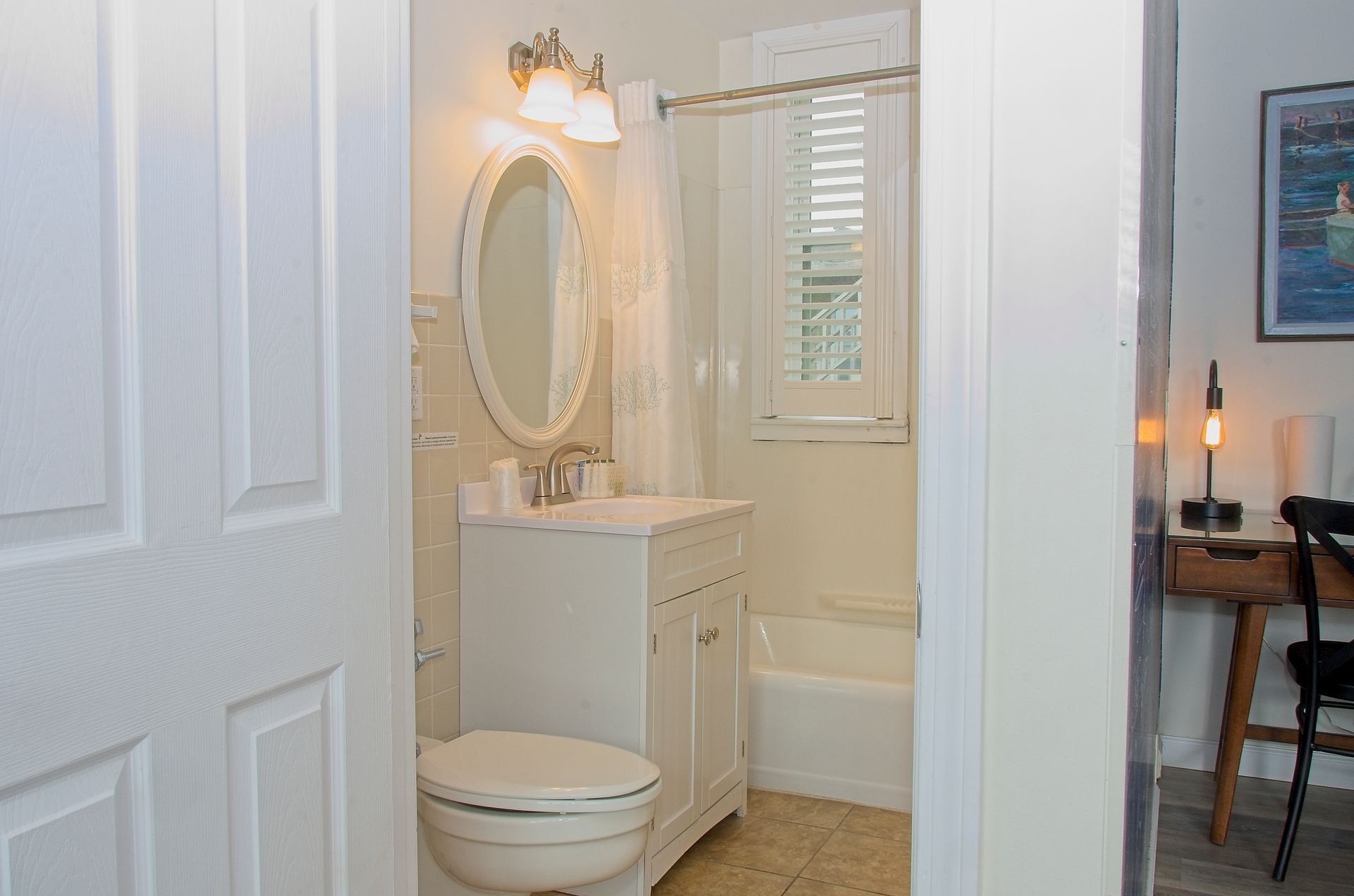 Bathroom with white vanity, oval mirror, and toilet. Doorway on the left leads into the space, with a desk visible on the right.