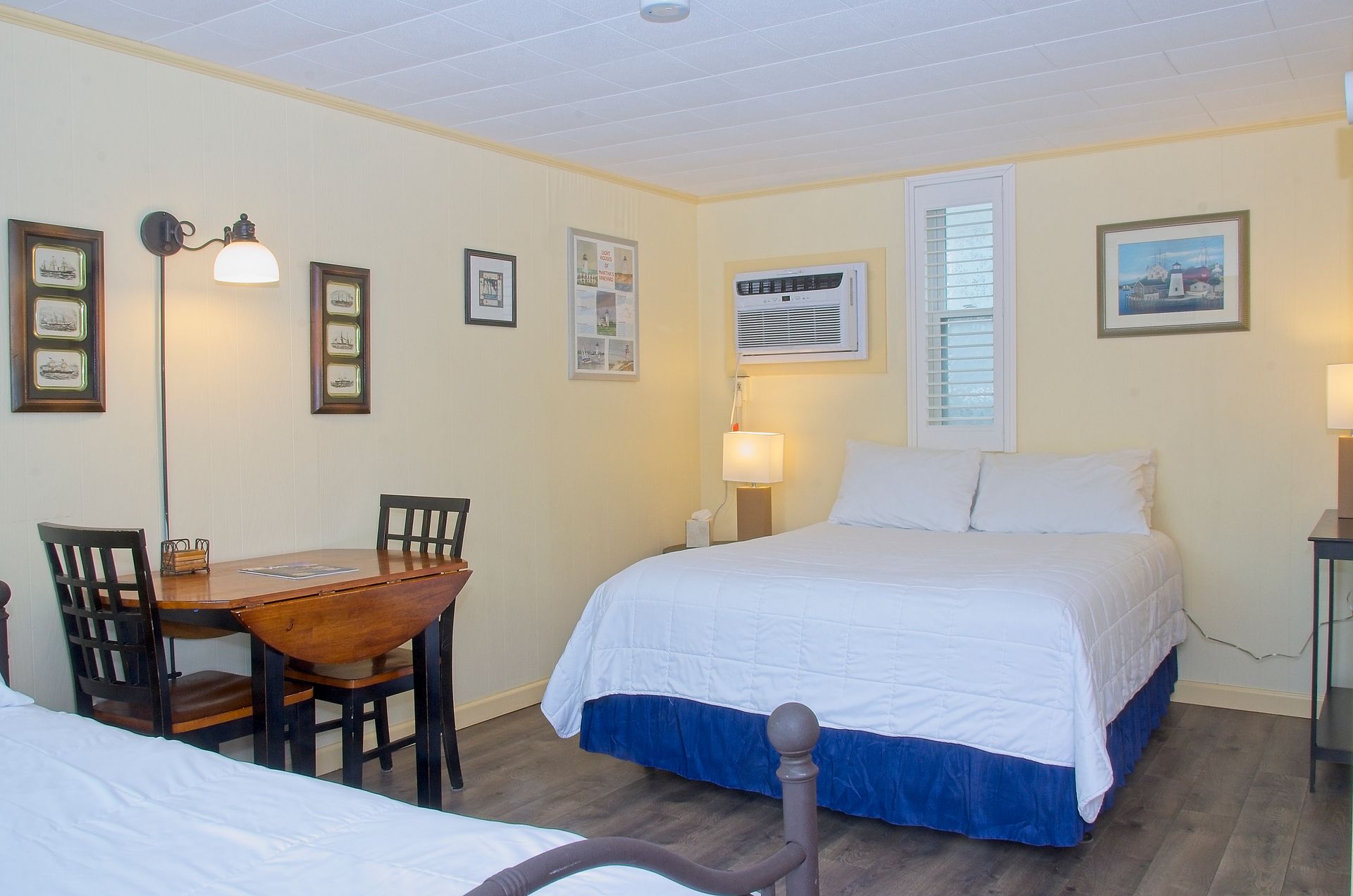 A small bedroom with two beds, a table with chairs, and various framed pictures on yellow walls.