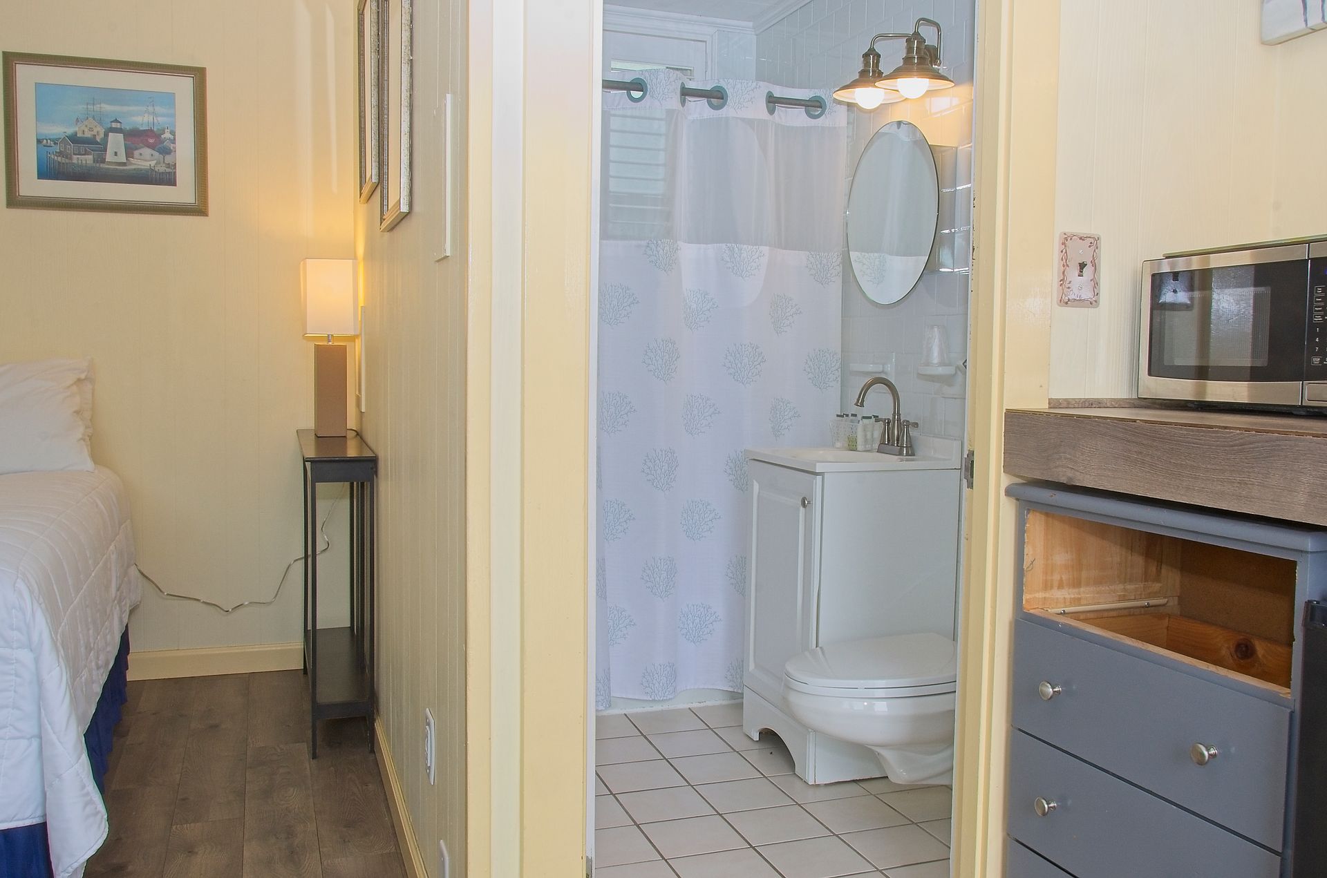 View of a small motel room interior with bathroom, bed, and microwave. The bathroom has a toilet and a small sink.