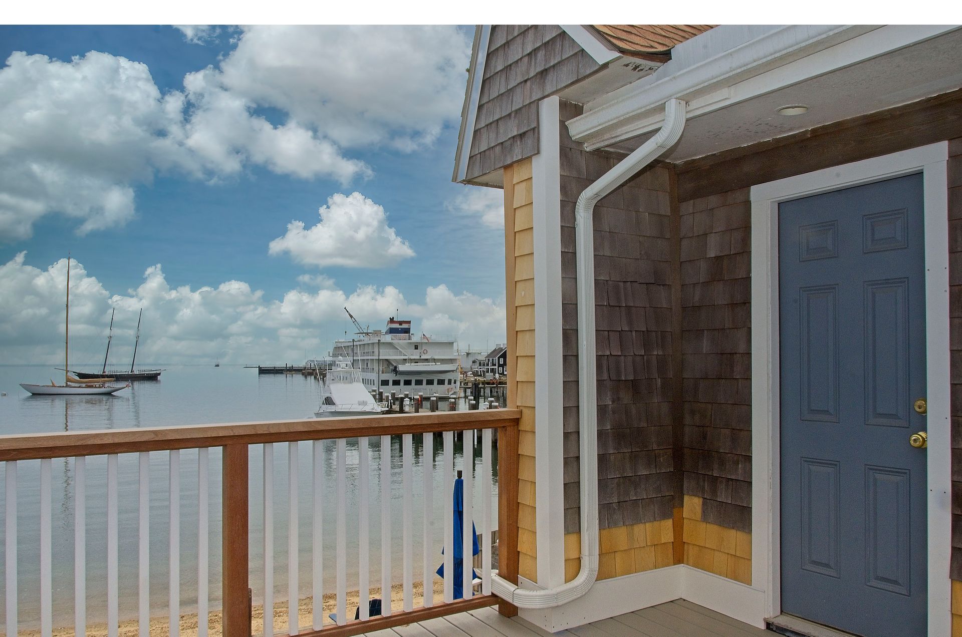 A seaside cottage with a blue door and a porch overlooking a harbor with boats and a pier under a cloudy sky.