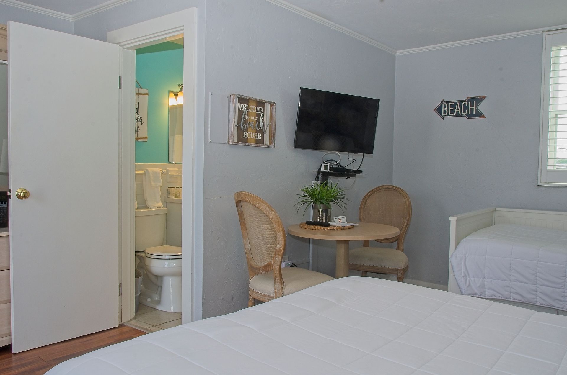 A small, light-filled hotel room with a queen bed, table, chairs, and a bathroom visible. A TV is mounted on the wall.