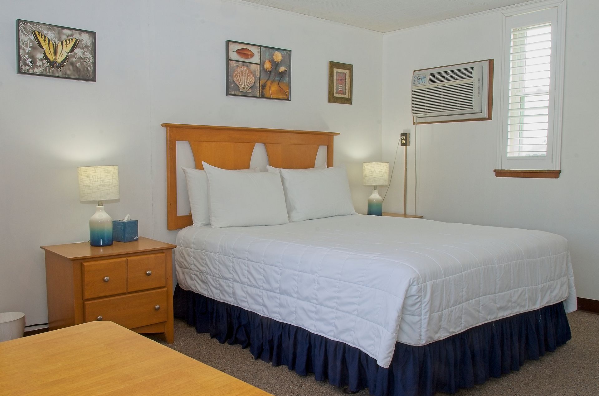 Bedroom with a queen-sized bed, nightstands, and wall art. Features white walls, a window-mounted AC unit, and a window with blinds.