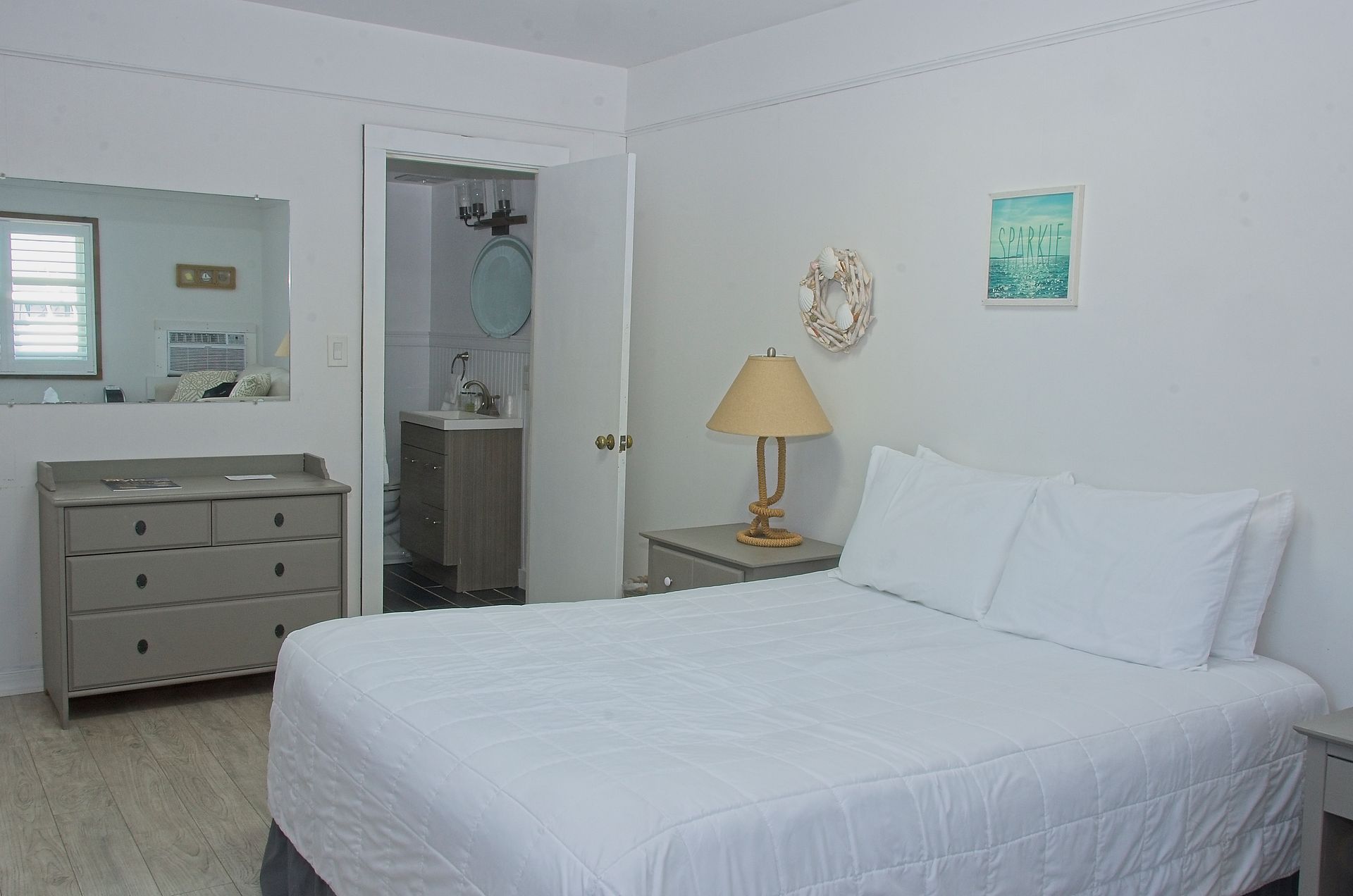 A white-walled bedroom with a bed, dresser, and an open doorway to a bathroom. The room is simply decorated with a muted color palette.