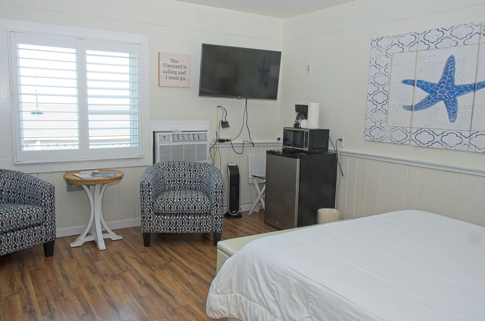 A bright, coastal-themed hotel room with a bed, two patterned armchairs, a window, and a flatscreen TV. A blue starfish decorates the wall.