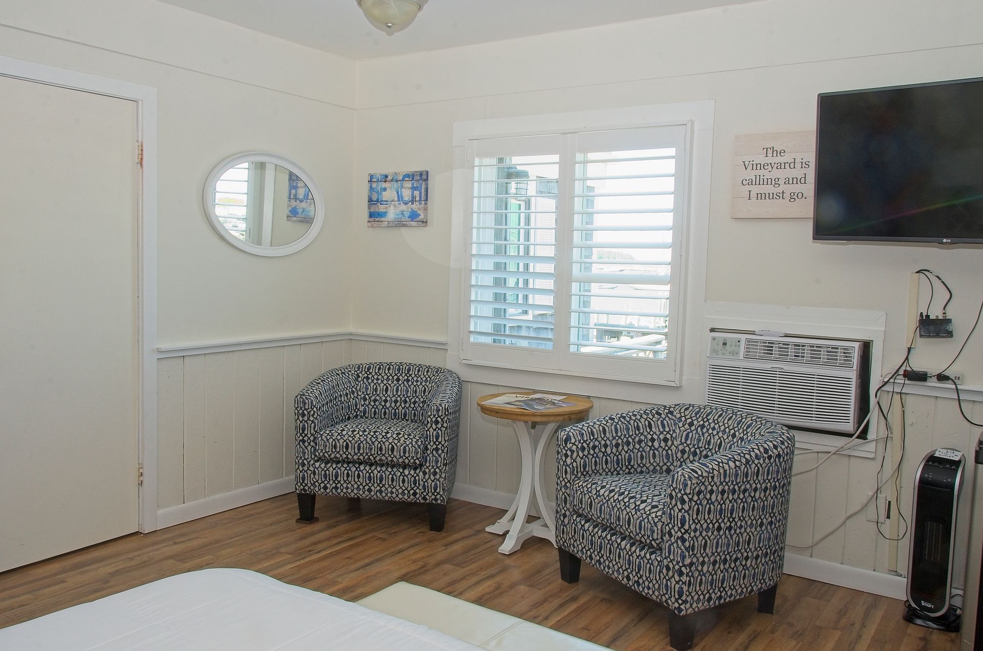 A small, cozy room with two patterned armchairs, a small table, and a window with shutters. A television is mounted on the wall.