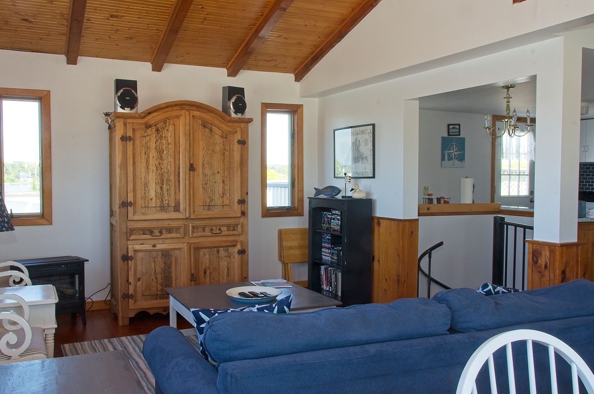 Living room with a large wooden cabinet, blue couch, and windows. A kitchen area is visible through a doorway.