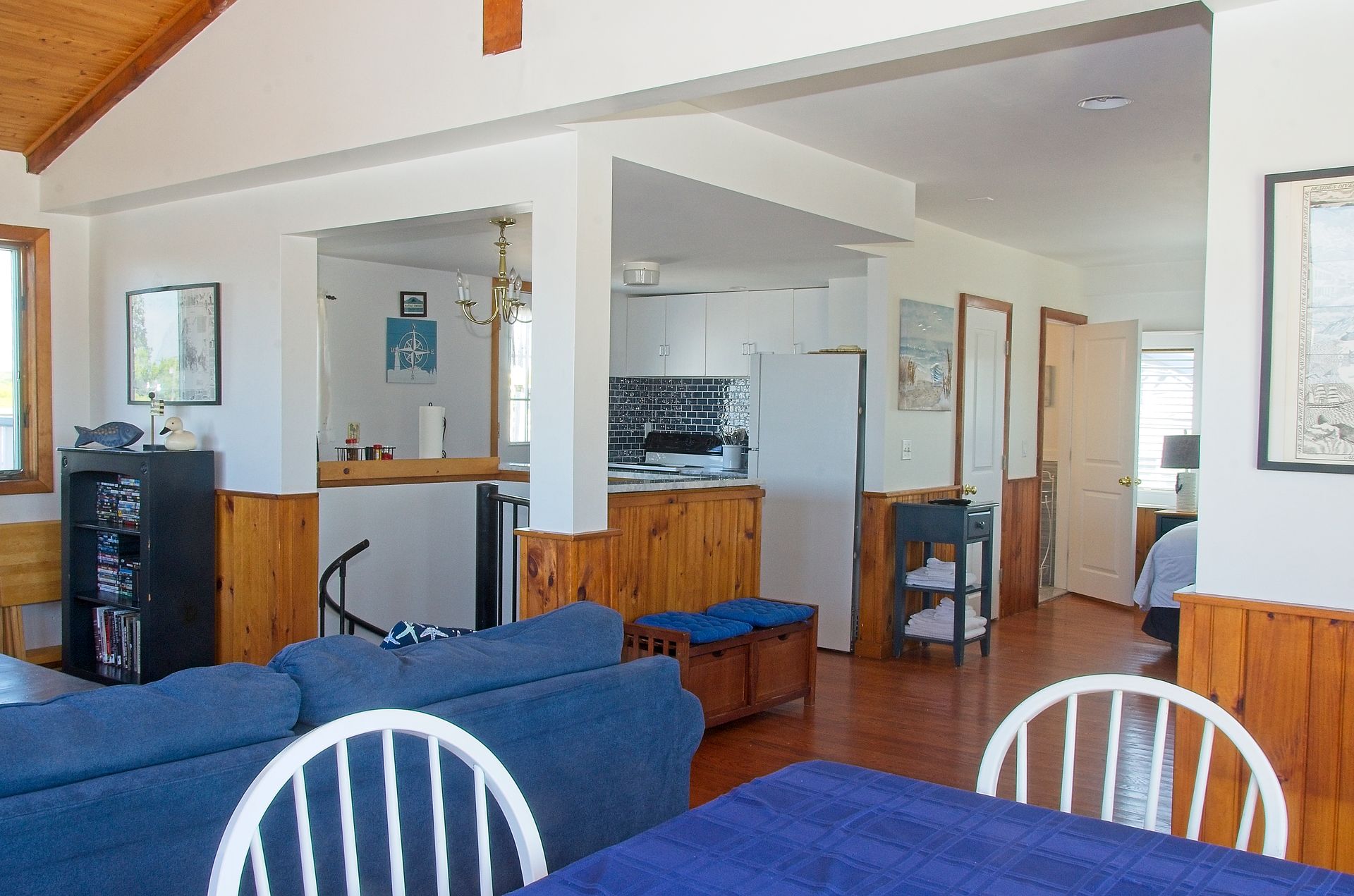Interior view of a cozy living space with blue sofa, wooden paneling, and open kitchen in the background. White chairs sit around a table.