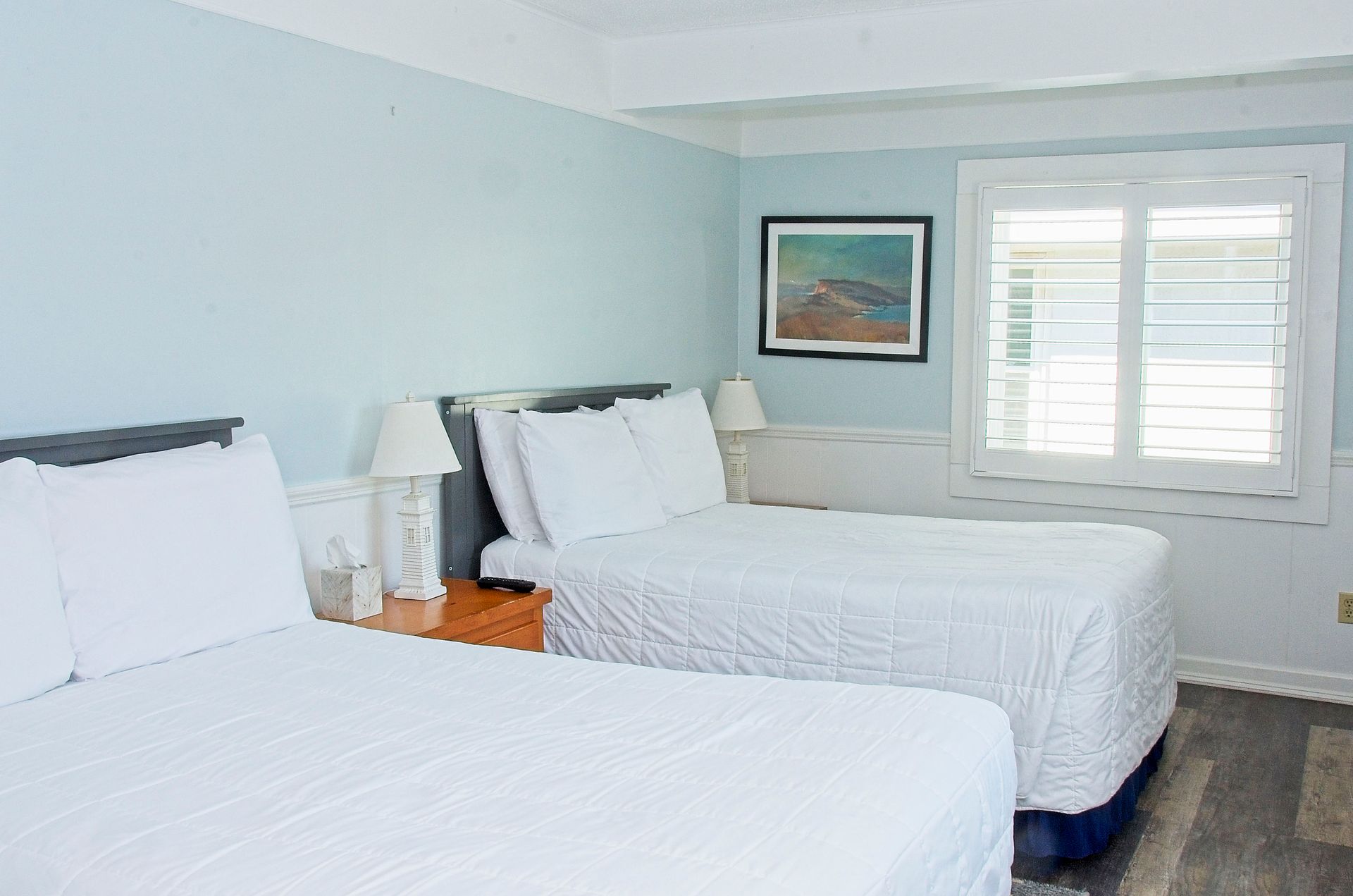 A hotel room with two beds, light blue walls, white bedding, and a window with shutters.