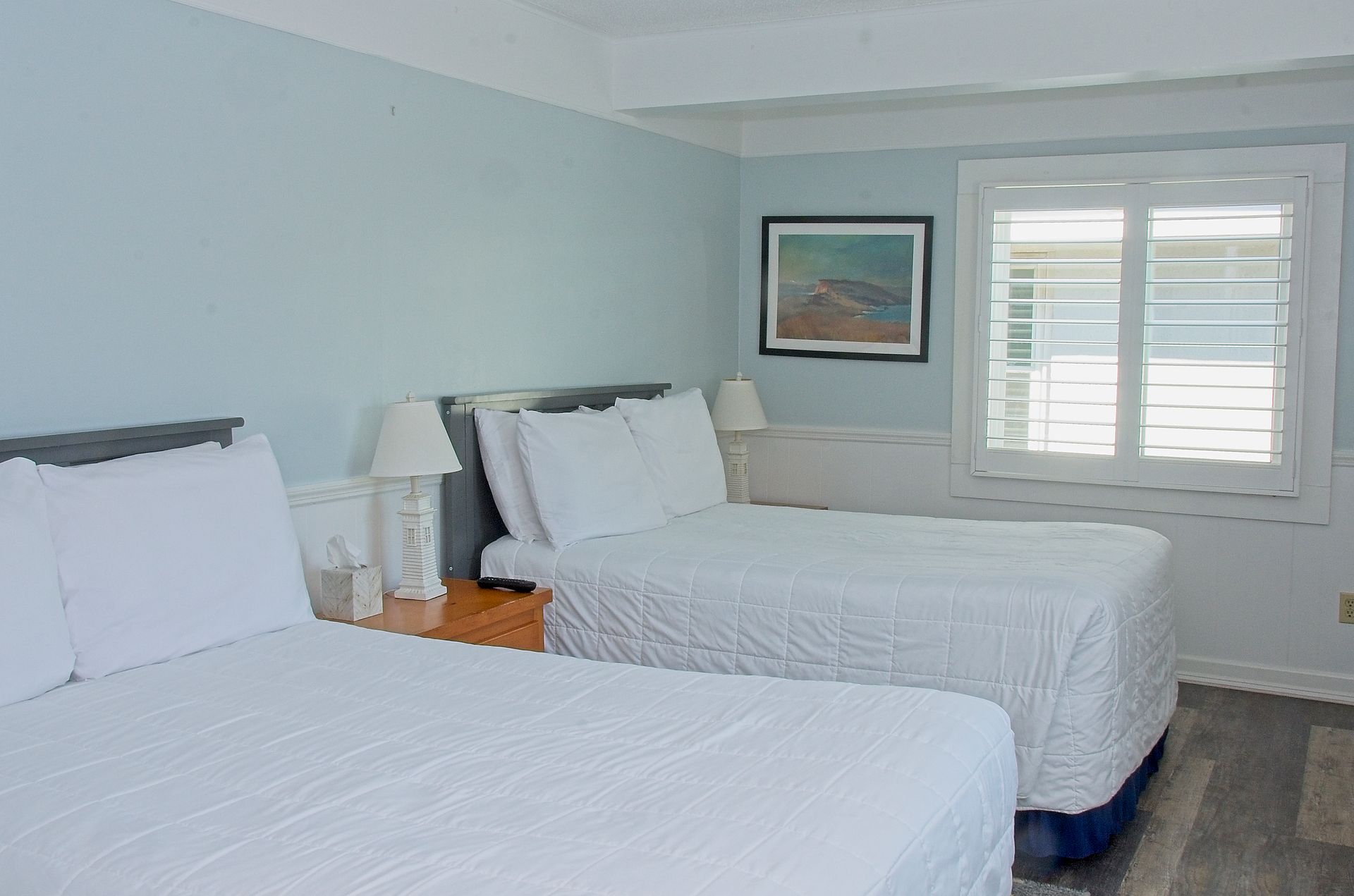 Two beds in a room painted light blue, with white bedding and shutters. A small artwork hangs above a radiator.
