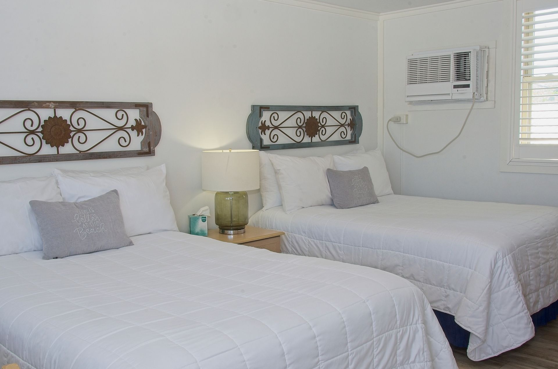 Two beds in a white-walled room, each with a white quilt and pillows. Decorative metal wall art hangs above.