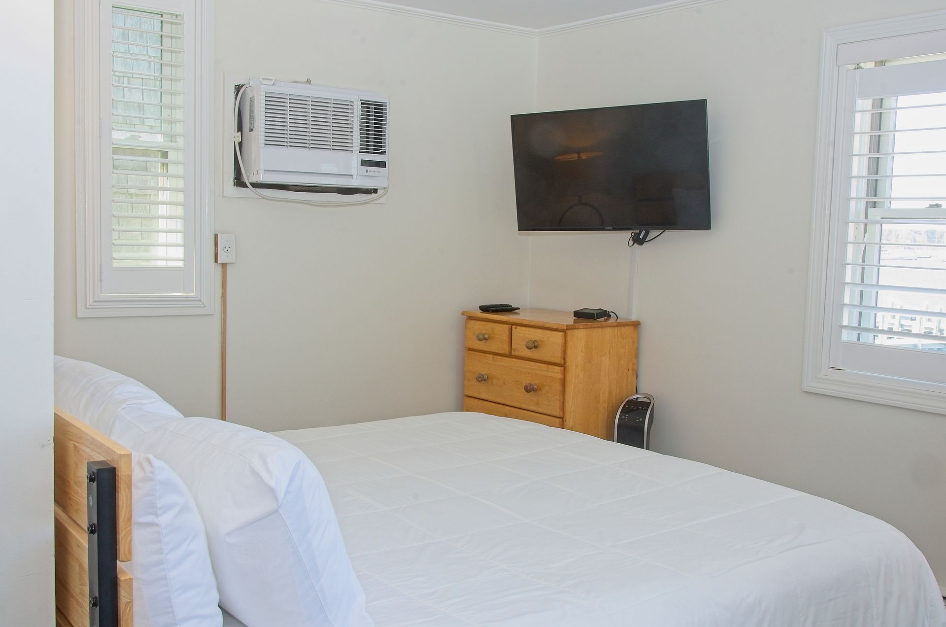 Bedroom with a white bed, wooden dresser, and a mounted TV. A window and air conditioning unit are also present.