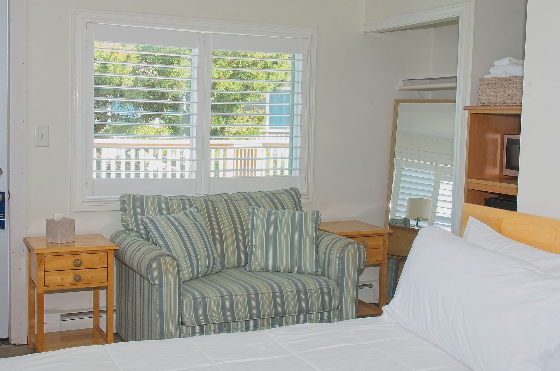 Bedroom interior with bed, striped loveseat, nightstands, window with shutters, and an open shelf with a microwave.