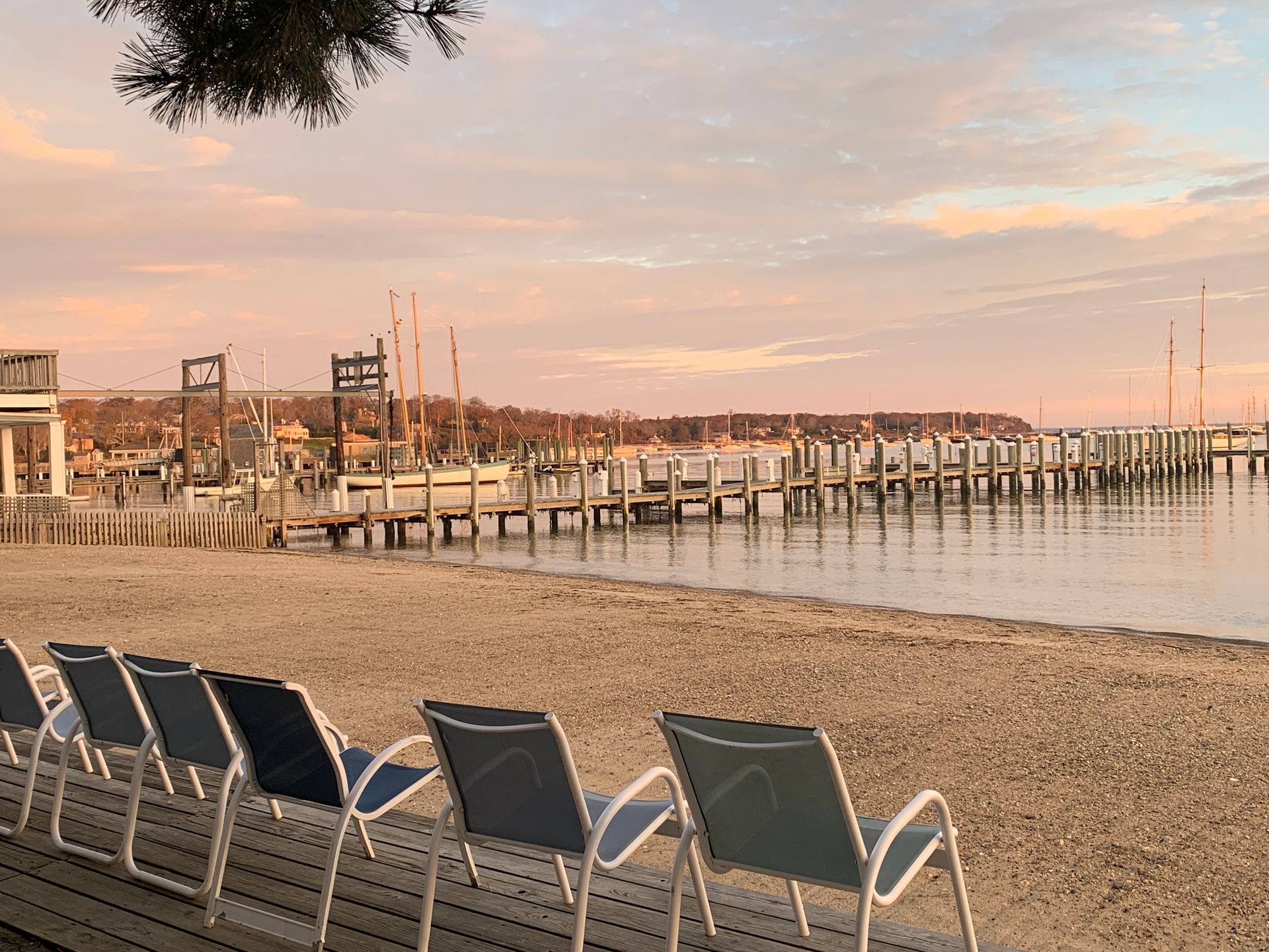 Beach with empty chairs facing calm water and docks. Pink and orange sunset colors the sky.