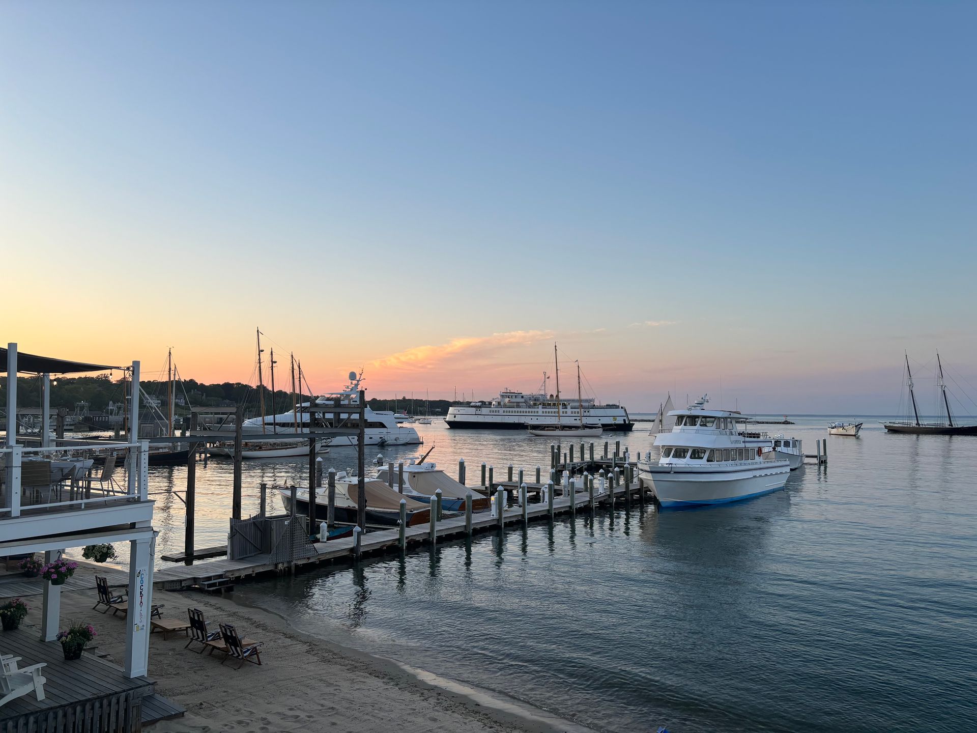 Boats docked at a harbor at sunset, with a clear blue and orange sky. Some people are on the docks and beach.